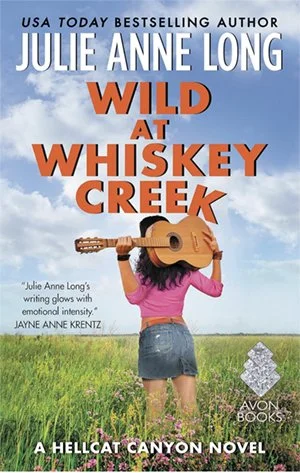 Book cover of 'Wild at Whiskey Creek' by Julie Anne Long, showing a woman in a pink shirt and denim shorts holding a guitar over her shoulder, standing in a grassy field with a blue sky background.