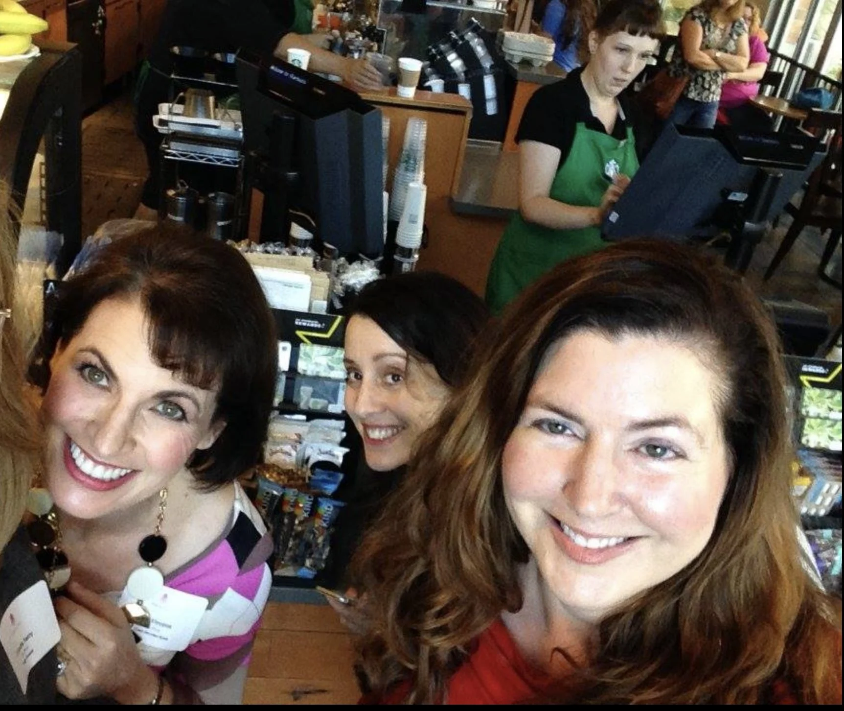 Three smiling women taking a selfie inside a coffee shop. Two women are in the foreground, one with dark brown hair and a red top, the other with dark hair and a colorful top. A third woman, with black hair, is in the background behind them.