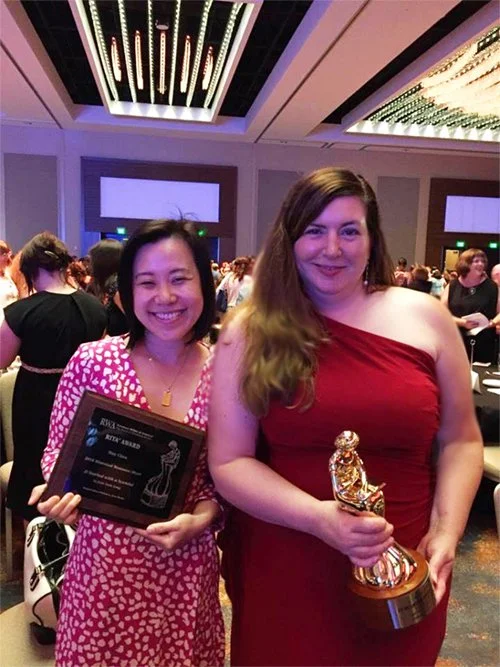Two women at an award ceremony holding trophies, smiling, in formal dresses, with a crowd in the background.