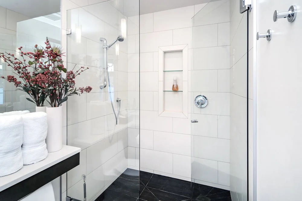 Modern bathroom with white tiled walls, a glass shower enclosure, and a built-in shower niche with a small bottle. A black shelf holds rolled white towels and a vase with pink flowers, and there are hooks on the door for hanging items.
