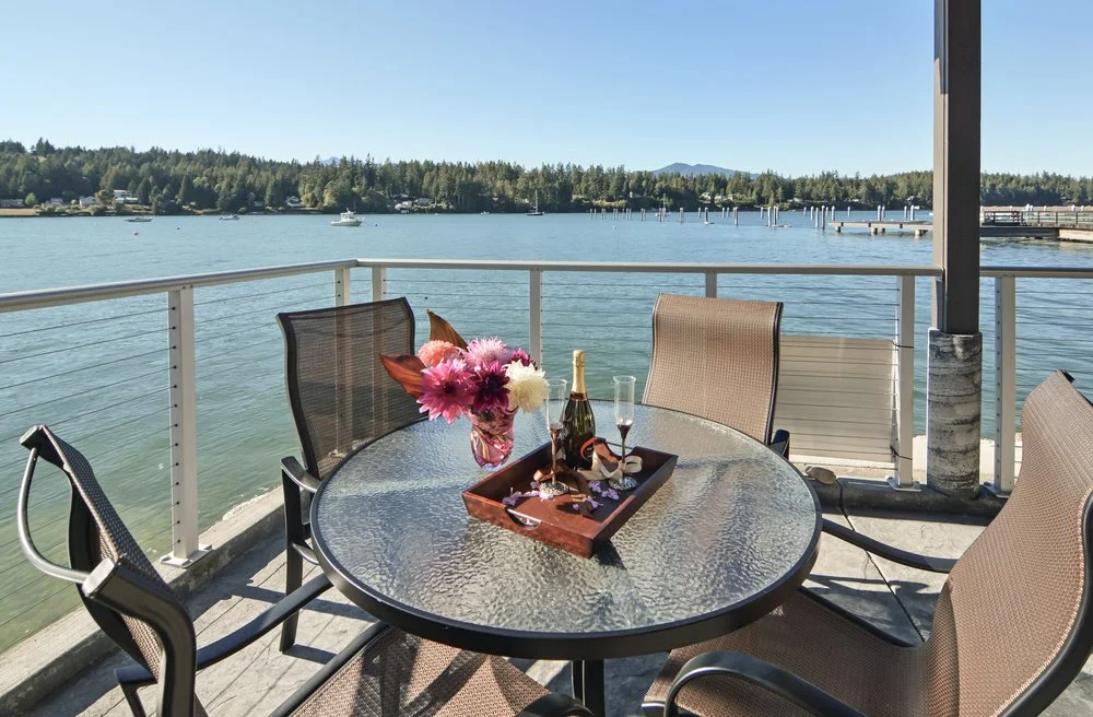 Outdoor patio table with a vase of pink and white flowers, a bottle of champagne, a glass, and jewelry, overlooking a lake with boats and docks under a clear blue sky.