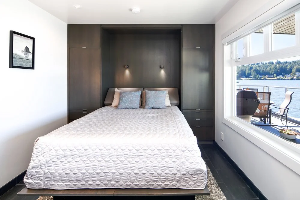 Bedroom with a large bed with pillows, dark built-in cabinetry, and a large window showing a view of water, trees, and outdoor seating.