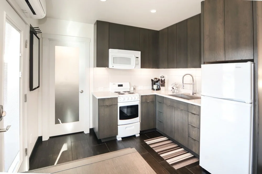 Modern kitchen with dark wood cabinets, white appliances including a refrigerator, microwave, and stove, a white sink with a gooseneck faucet, and a striped rug on dark hardwood floor.