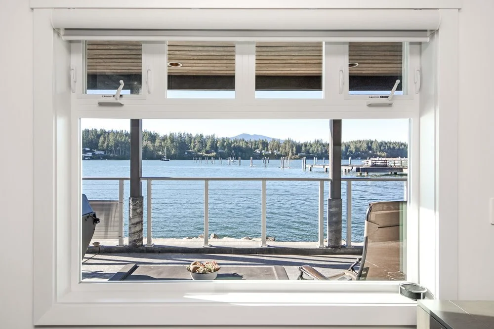 View of a calm body of water with a dock, outdoor seating, and trees in the background through a large window from inside a house.