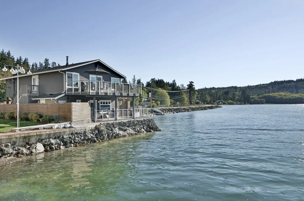 A house by a body of water with a rocky shoreline, surrounded by trees on a sunny day.