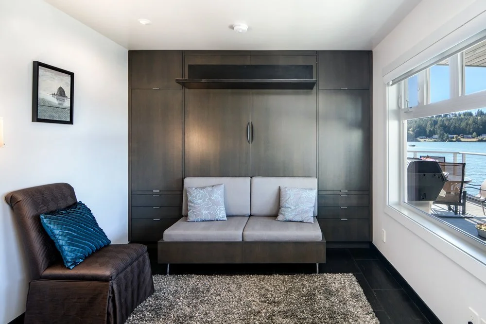 Living room with a beige loveseat, dark wood cabinets behind it, a brown armchair with a blue pillow, a framed picture on the wall, a window showing a waterfront view, and a carpet on the floor.