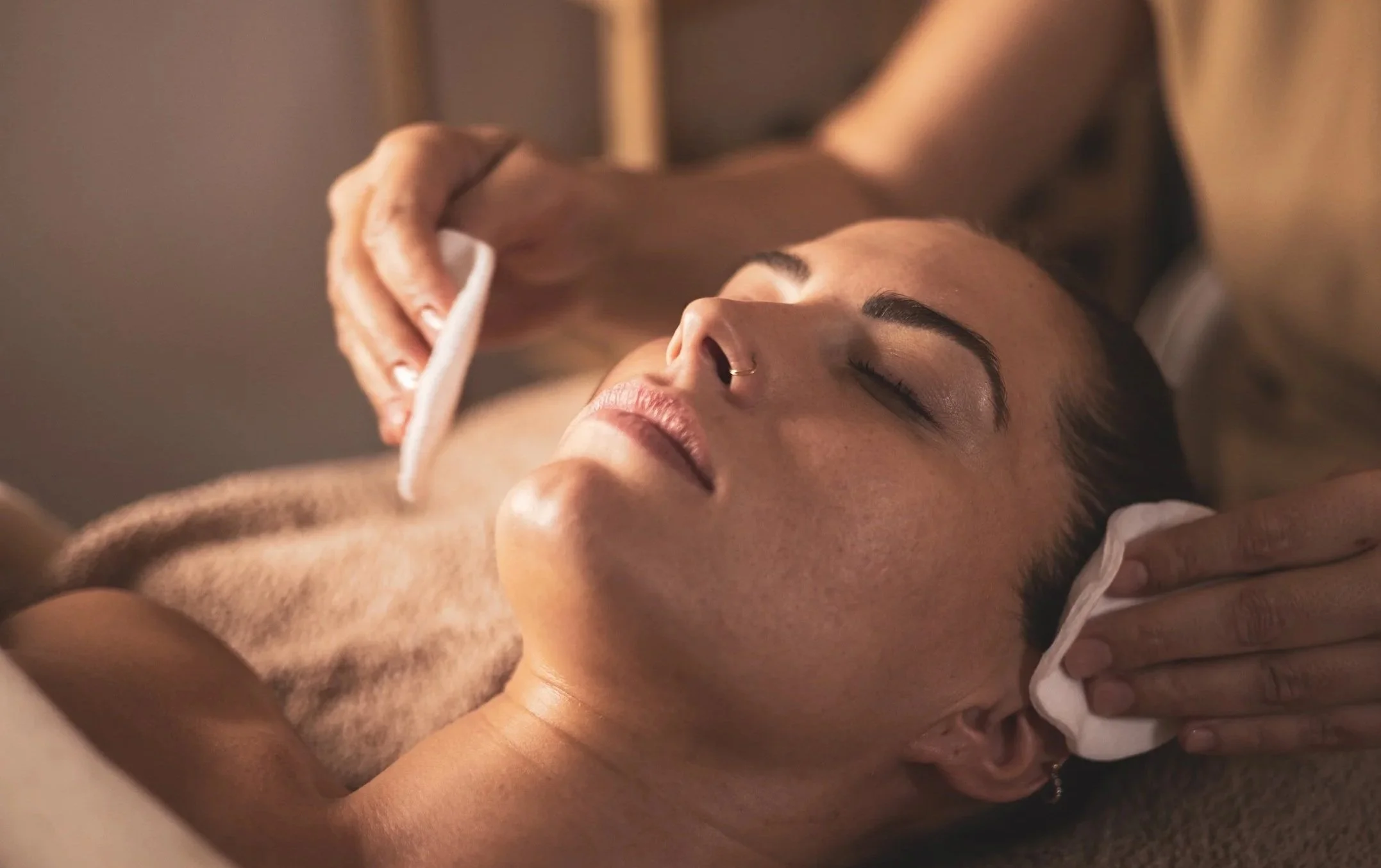 A woman lying down with eyes closed while a person gently wipes her face with a cotton pad.