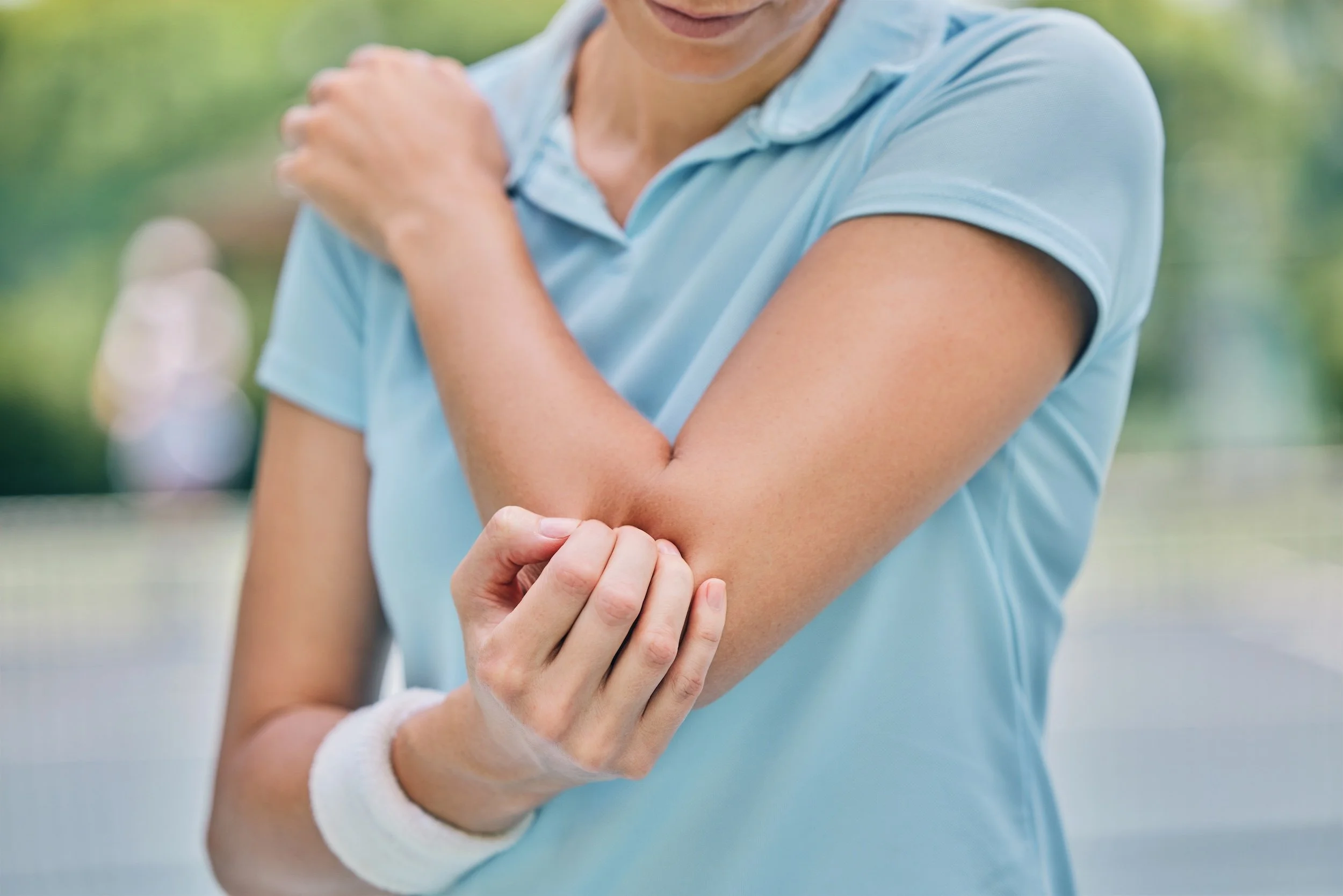 Close-up of a woman in a light blue shirt scratching her arm, with a white wristband, in an outdoor setting with blurred green background.