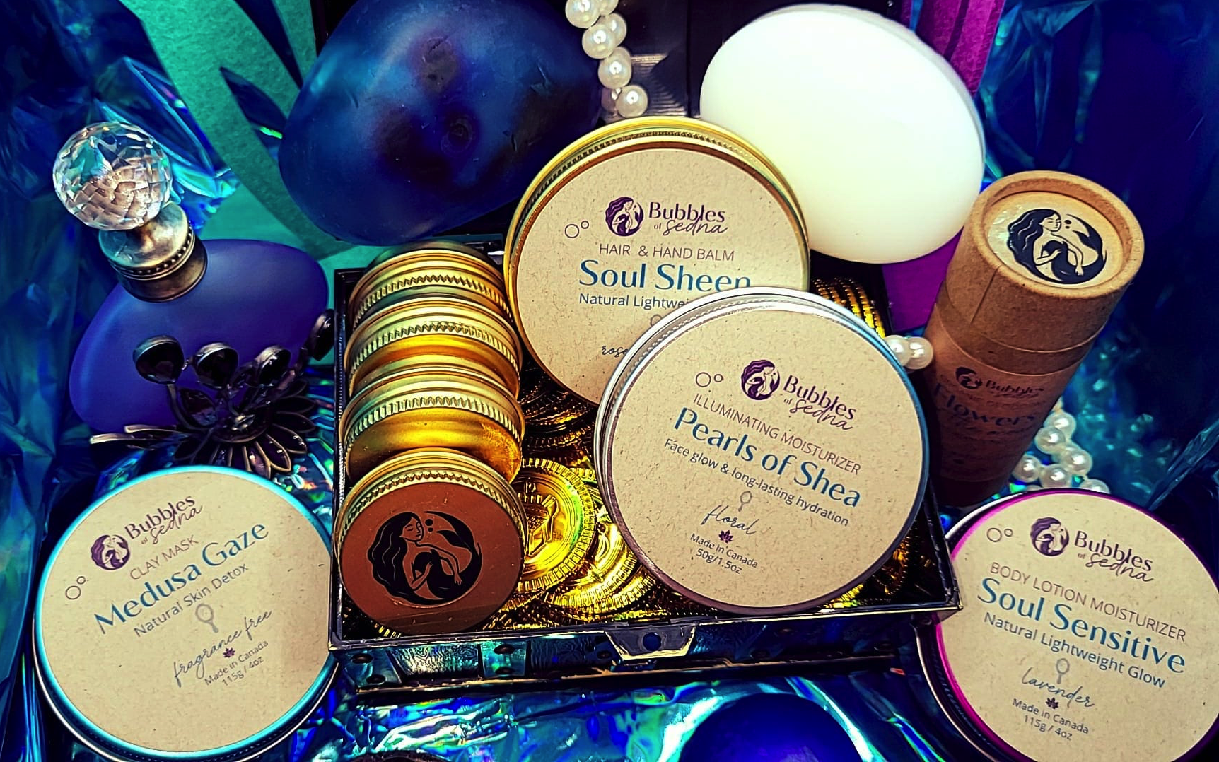 A collection of skincare and beauty products including jars of Bubbles of Sedna Soul Sheen balm, Pearls of Shea moisturizer, Medusa Gaze clay mask, a small container of flower-shaped body lotion, a decorative bottle, and nearby jewelry with pearls, set on colorful foil and cloth.