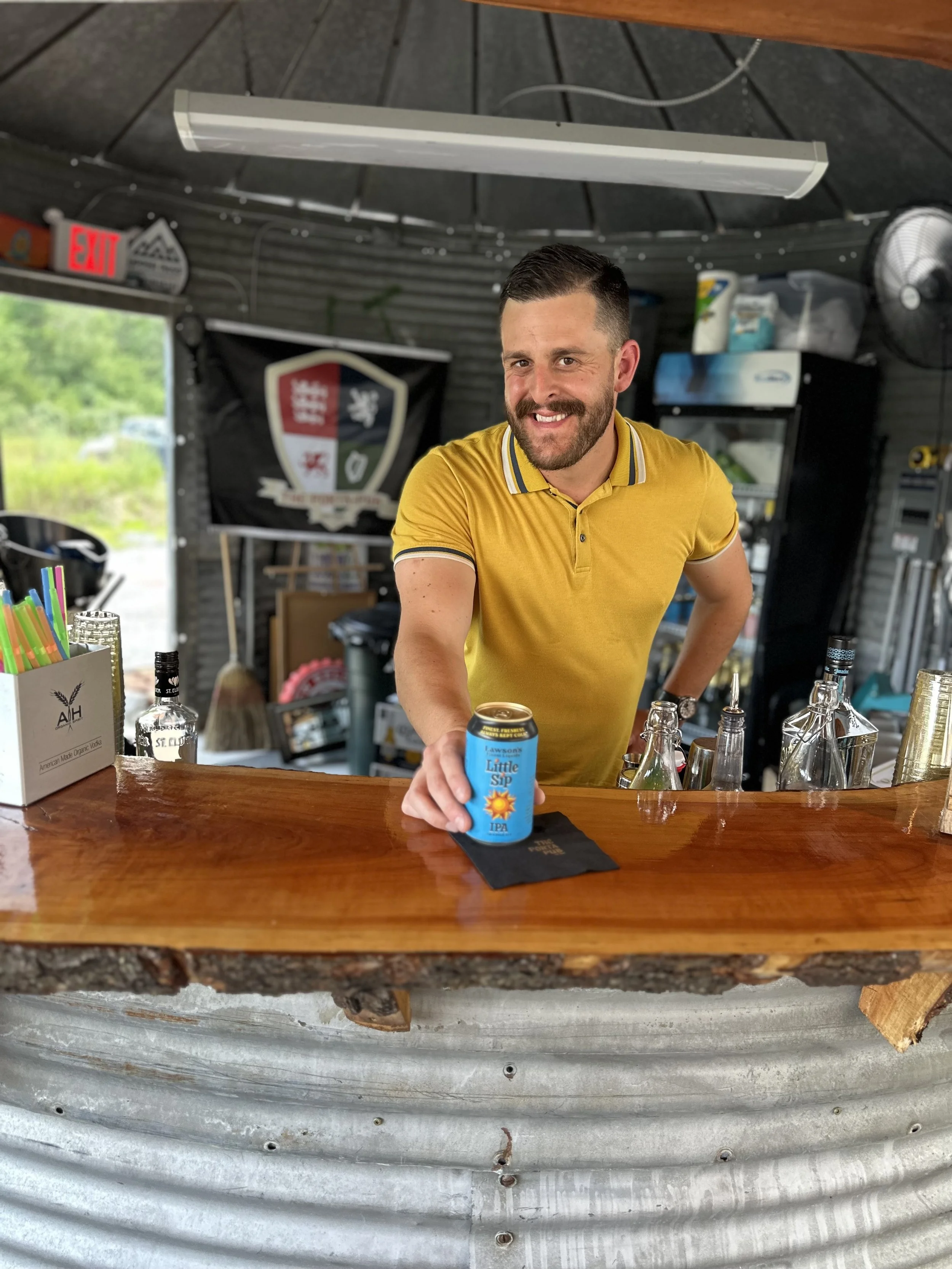 A smiling man in a yellow polo shirt standing behind a bar counter, holding a canned drink with the label 'Little Sip IPA,' in an outdoor setting with a rustic bar and various bottles and bar tools behind him.