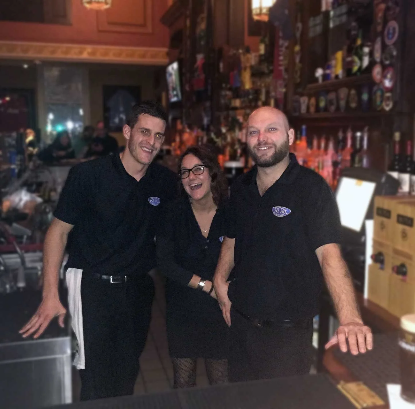 Three people in a bar, two men and one woman, smiling and posing for the photo. They are wearing black shirts with a logo that says 'RIRA'.