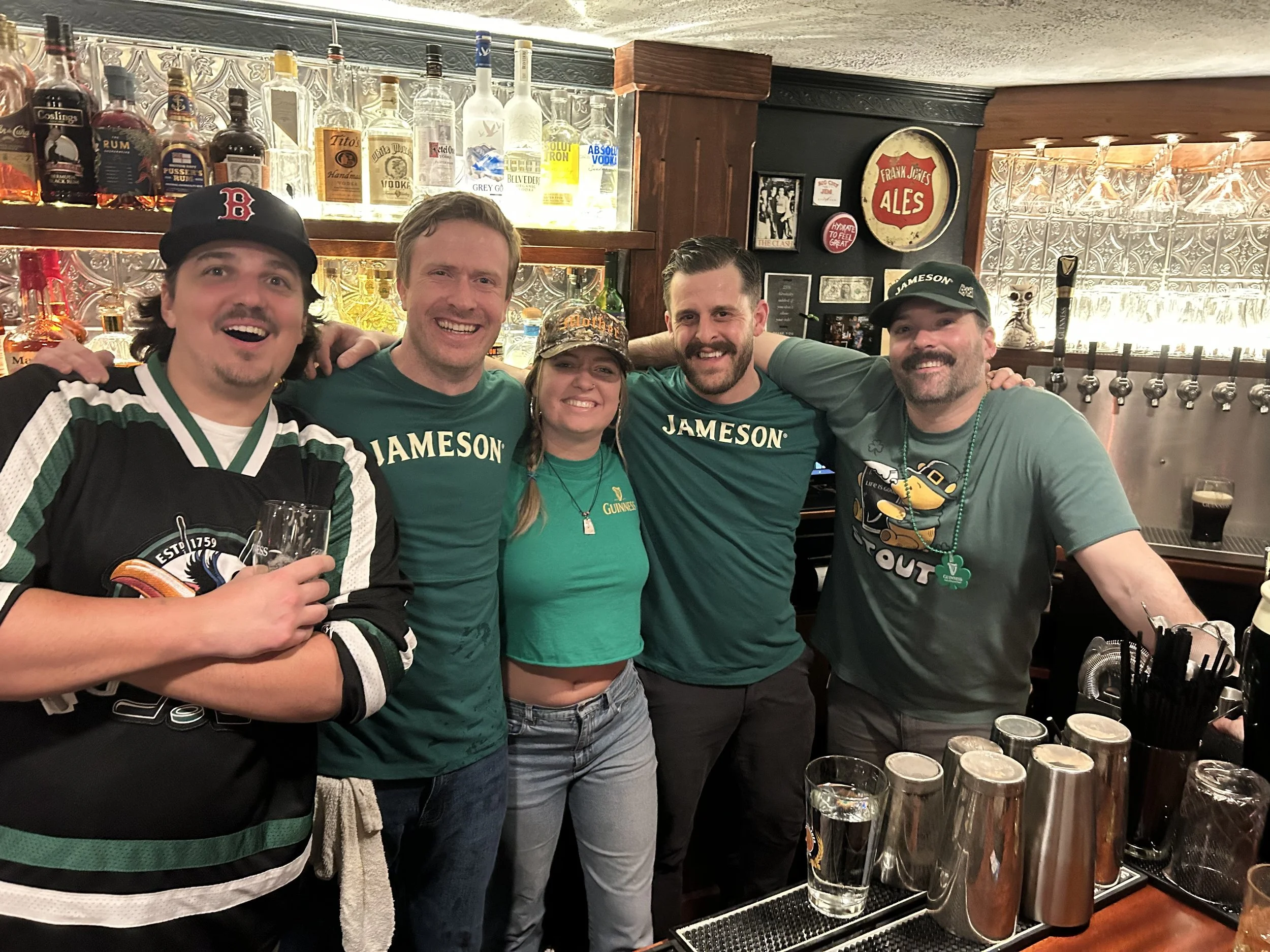 Group of five friends smiling at a bar, with liquor bottles on shelves behind them, celebrating St. Patrick's Day, wearing green and patriotic shirts.