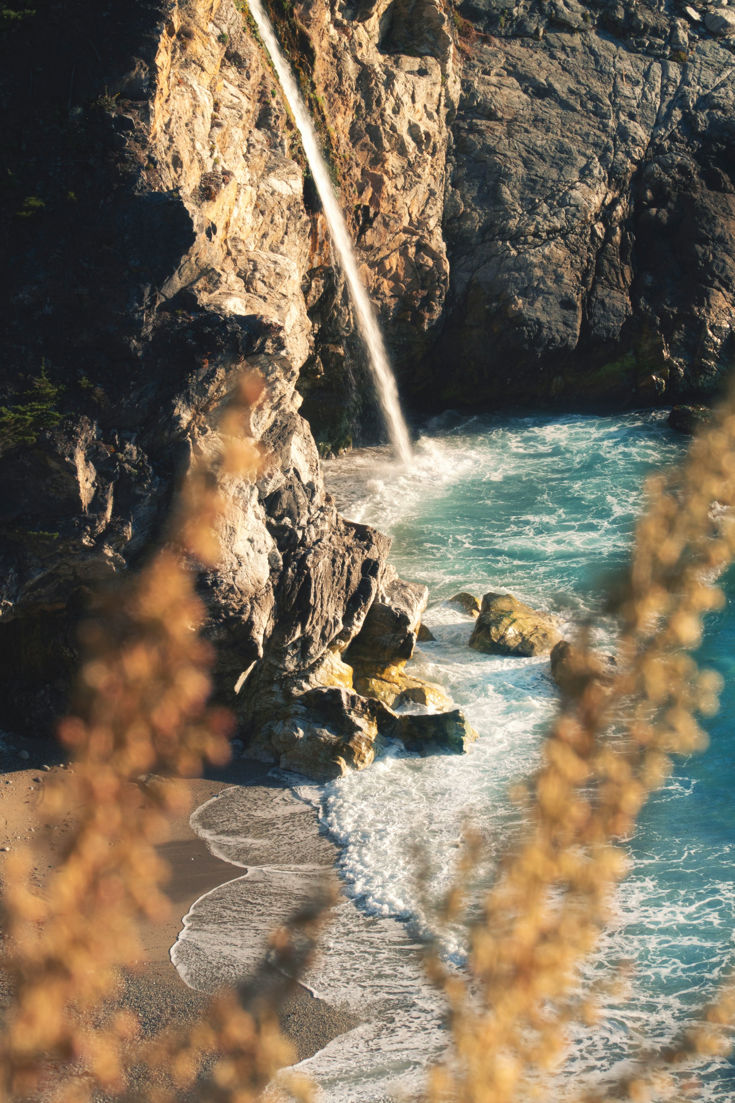 A scenic coastal scene with a waterfall flowing down rocks into the ocean, with brownish plants in the foreground.