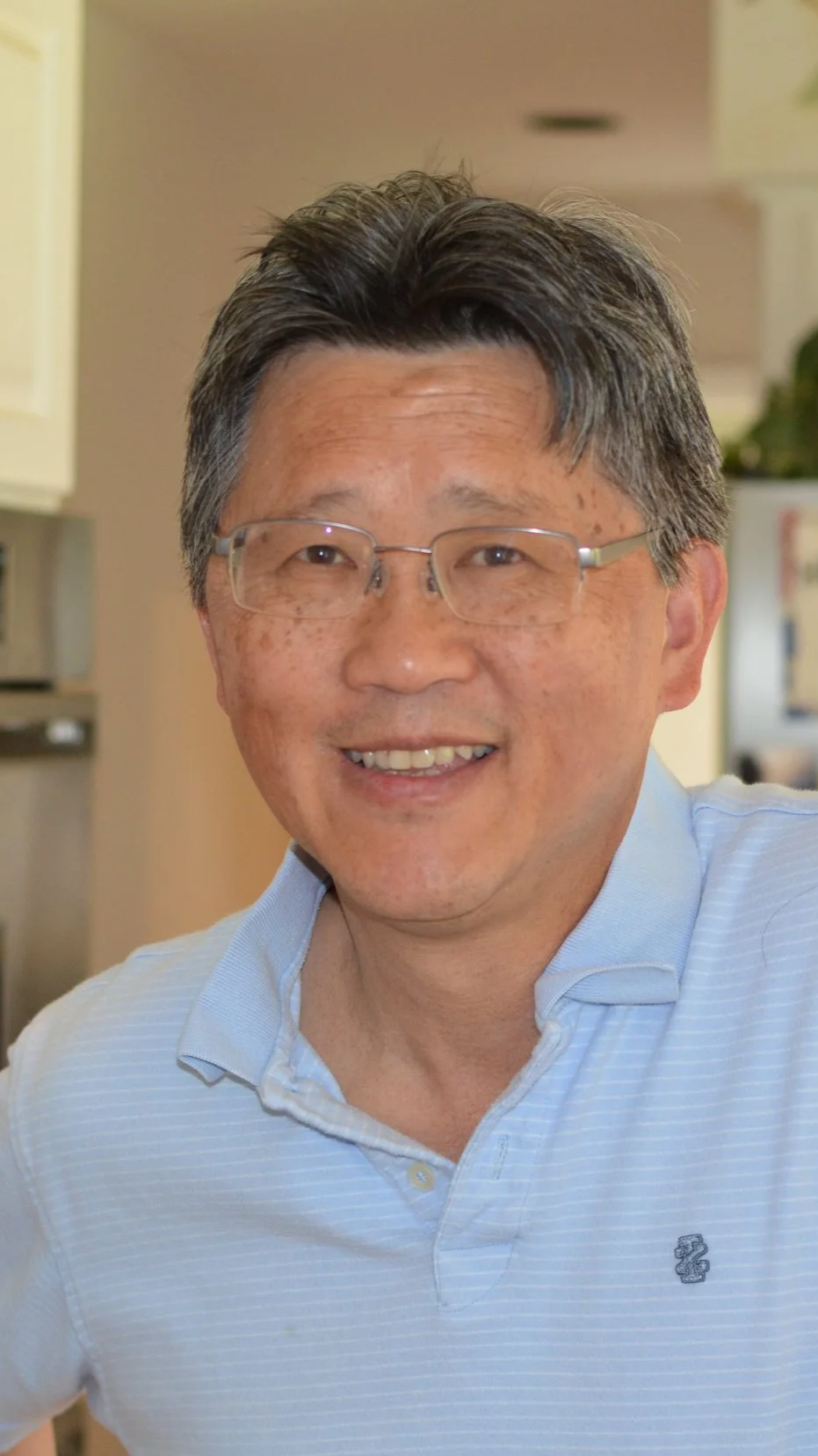 A smiling man with glasses, dark hair with gray streaks, wearing a light blue collared shirt, in an indoor setting.