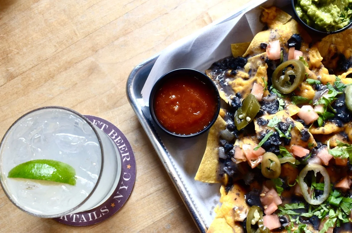 Plate of nachos topped with jalapeños, black beans, chopped tomatoes, and cilantro, with two cups of red salsa and a glass of clear soda with a lime wedge on a wooden table.