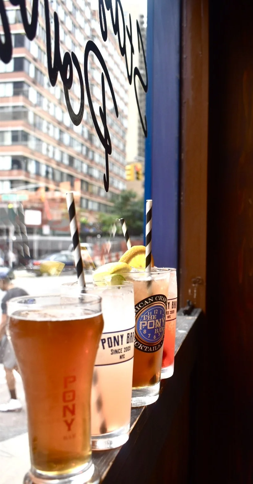 Several colorful cocktails with lemon and lime garnishes, lined up on a windowsill inside a bar, with a city street and buildings visible outside.