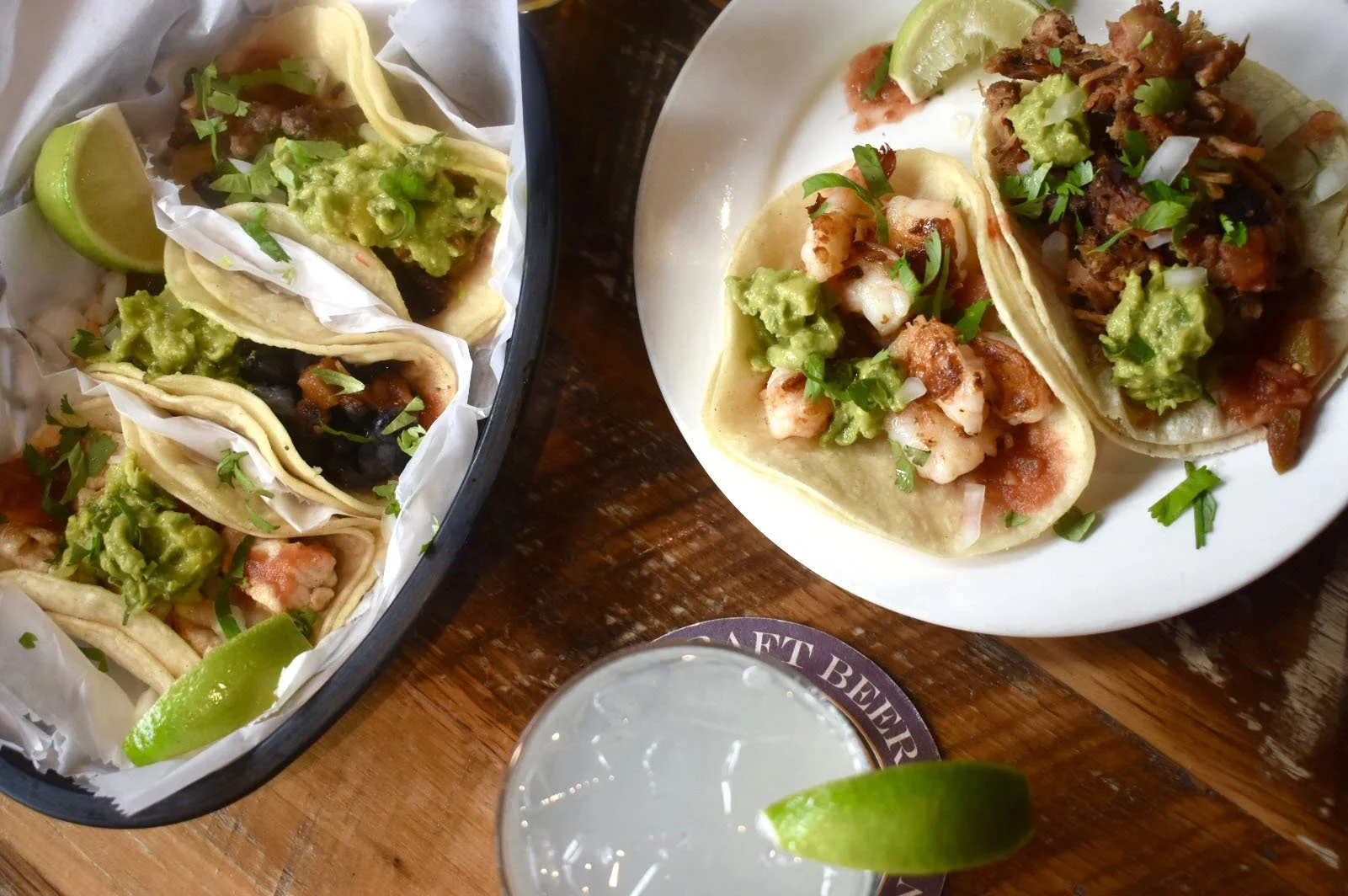 Three tacos filled with shrimp, guacamole, and onions on a white plate, with a side of lime wedges, a glass of beer with lime, and a tray of more tacos on a wooden table.