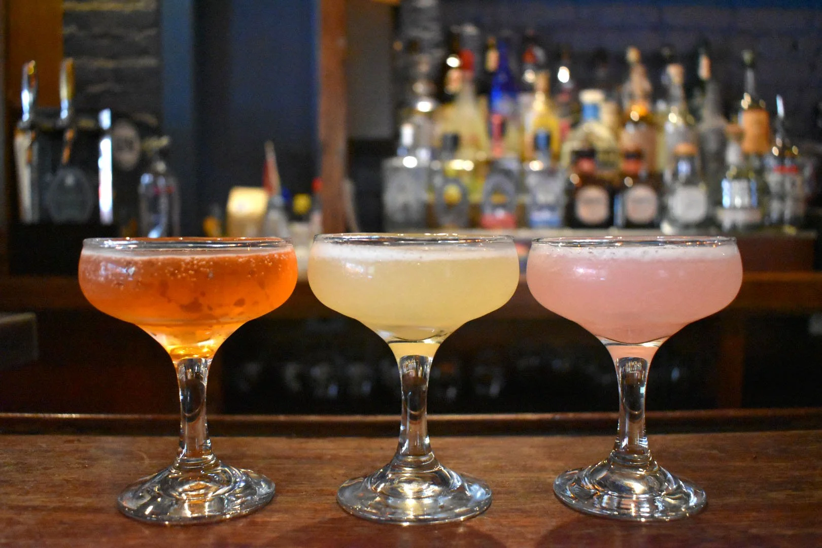Three colorful cocktails in coupe glasses on a bar counter with a blurred background of liquor bottles.