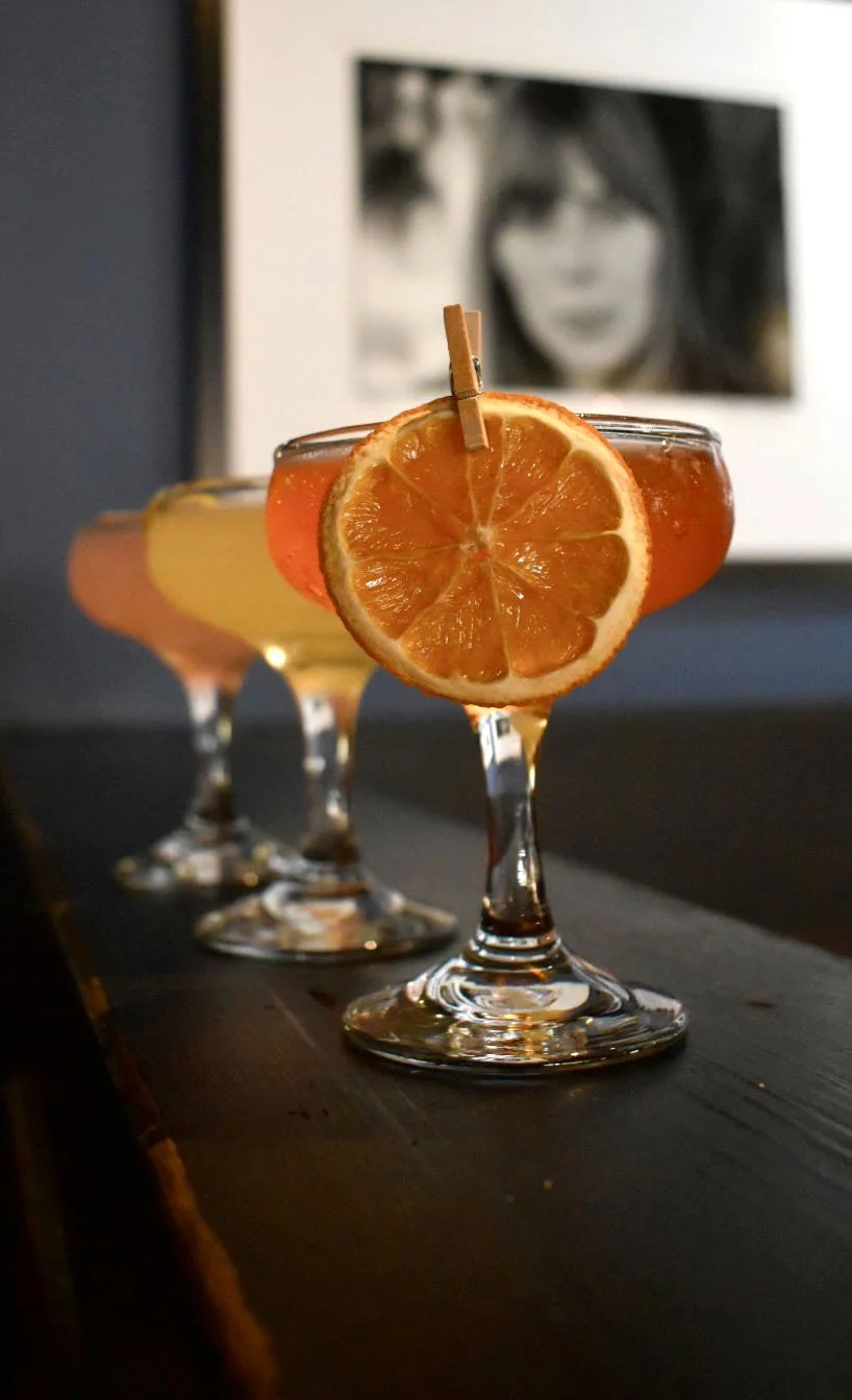 Three cocktails in coupe glasses garnished with orange slices and small clothespins, placed on a dark wooden surface with black-and-white framed photos in the background.