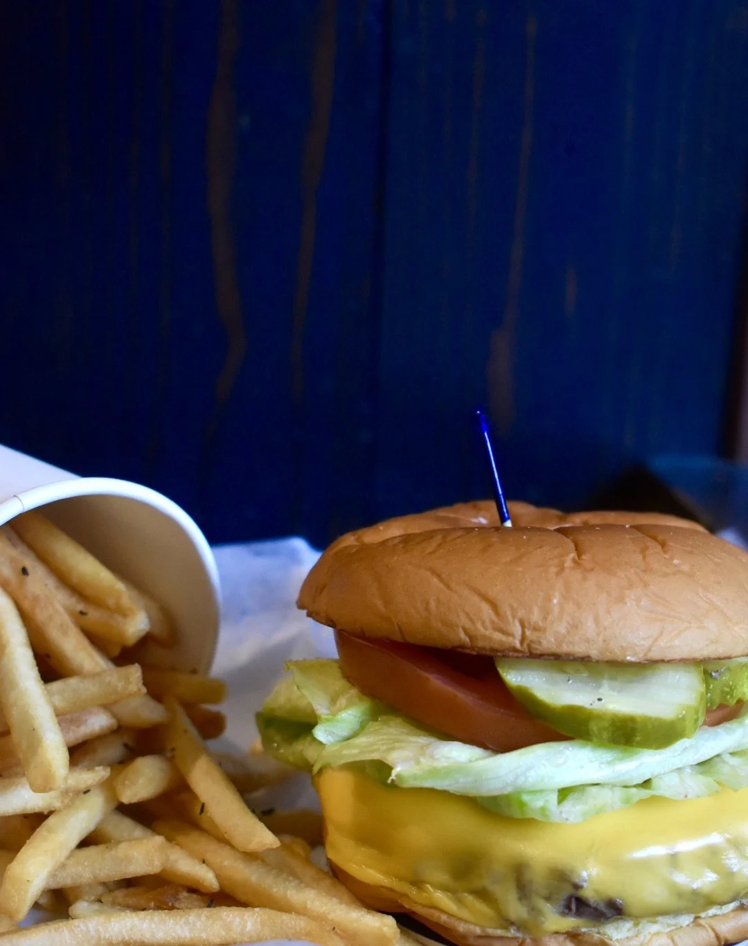 Cheeseburger with lettuce, tomato, pickles, and cheddar cheese, served with French fries in a white paper container.