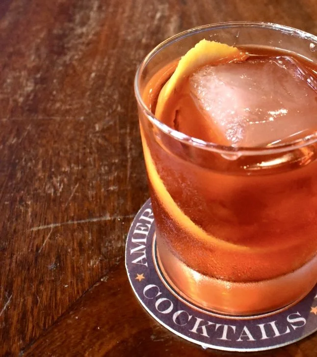 A glass of cocktail with an ice cube and a lemon peel on a coaster with 'Cocktails' written on it, placed on a wooden surface.