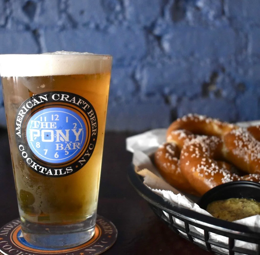 A pint of beer in a glass with a logo from The Pony Bar, a basket of pretzels, and a small dish of mustard on a dark table with a blue brick wall in the background.