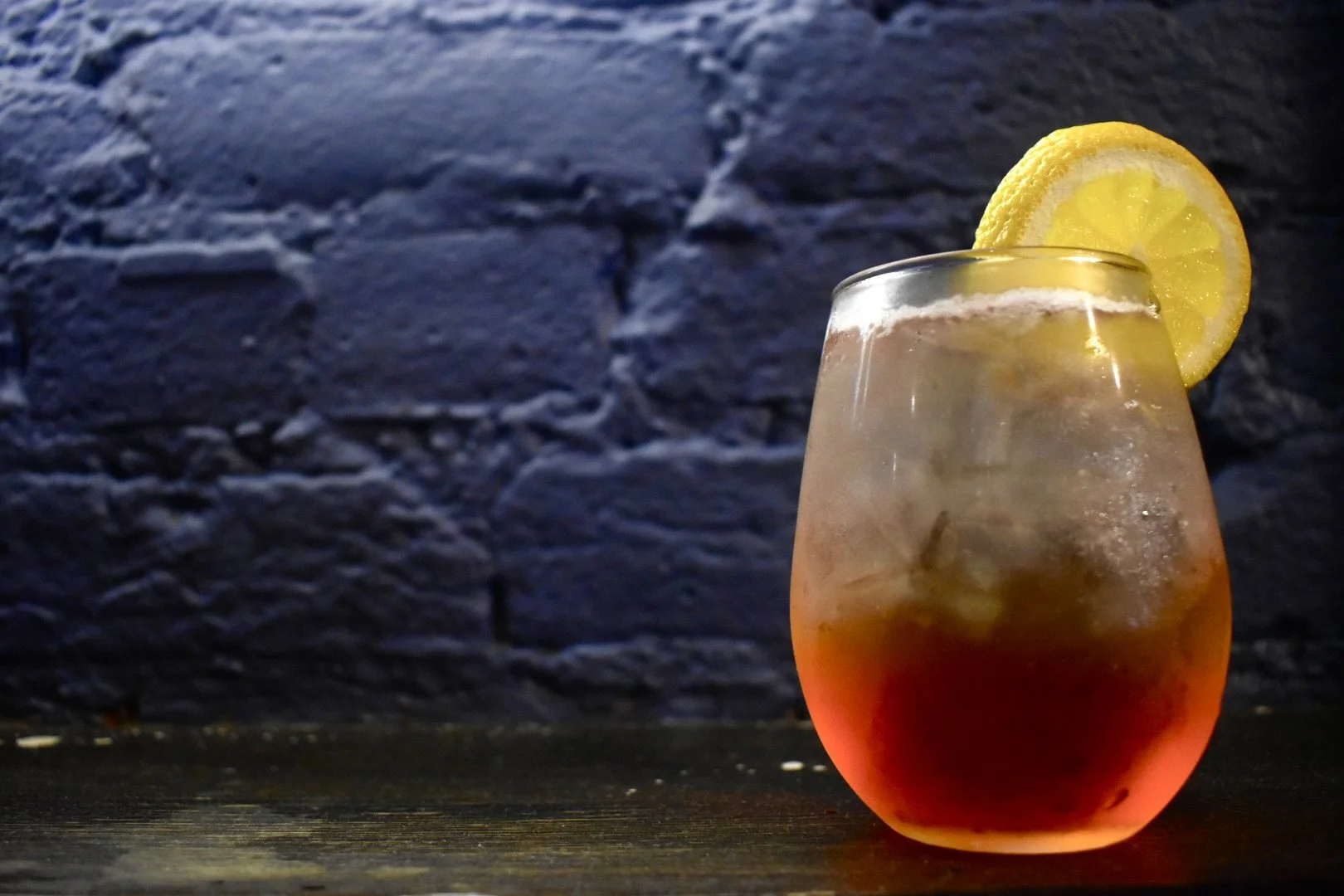 A glass of iced tea with a lemon slice on the rim, set against a dark brick wall background.