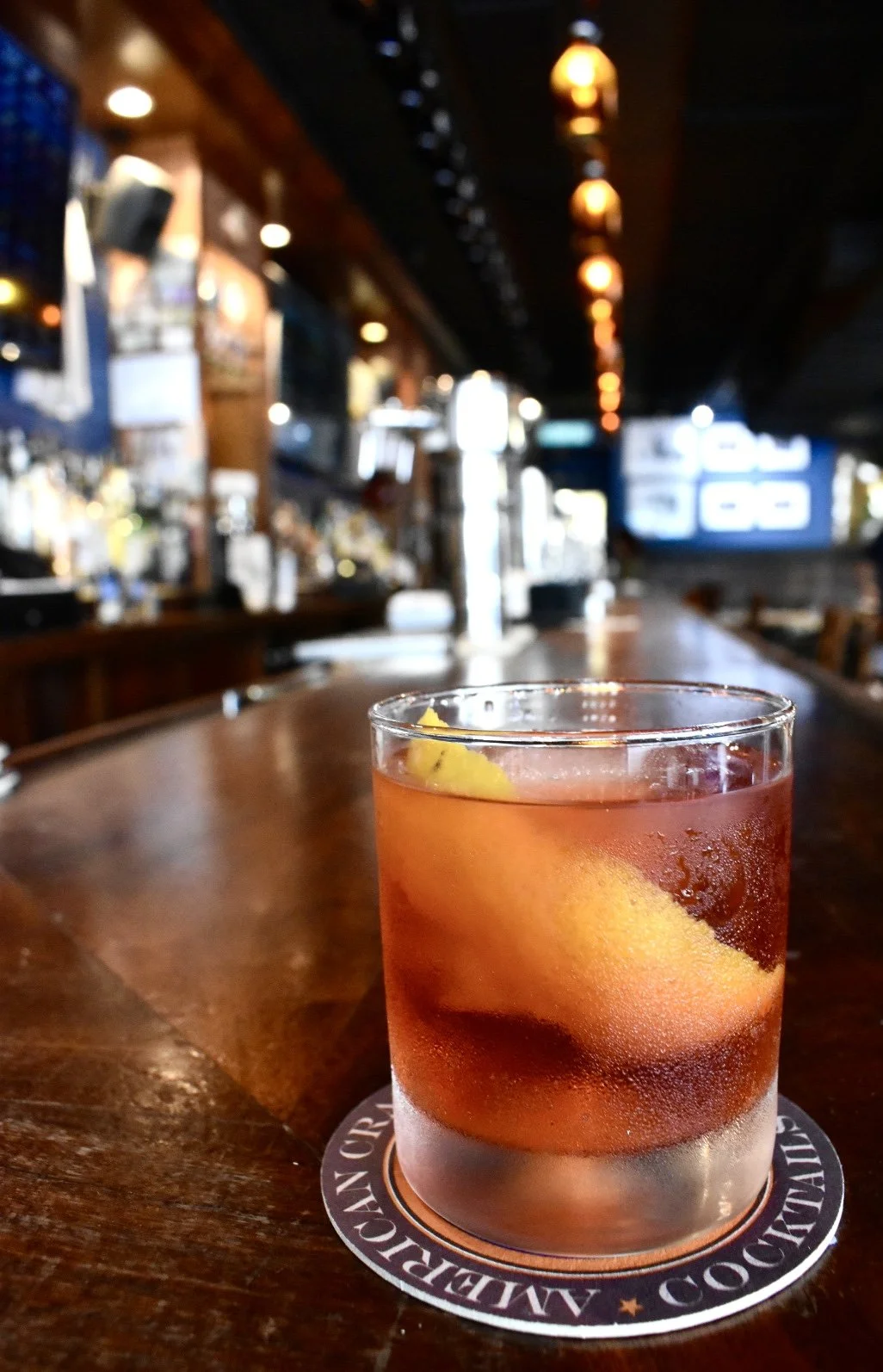 A glass of cocktail with a lemon twist on a wooden bar counter, with a blurry bar background and hanging lights.