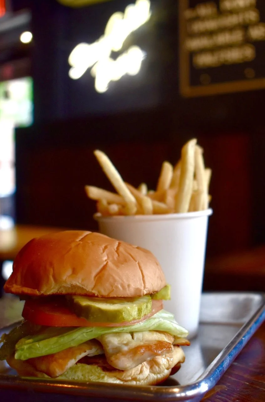 A sandwich with lettuce, tomato, pickles, chicken, and a bun served with a side of French fries in a white cup, on a metal tray at a restaurant.