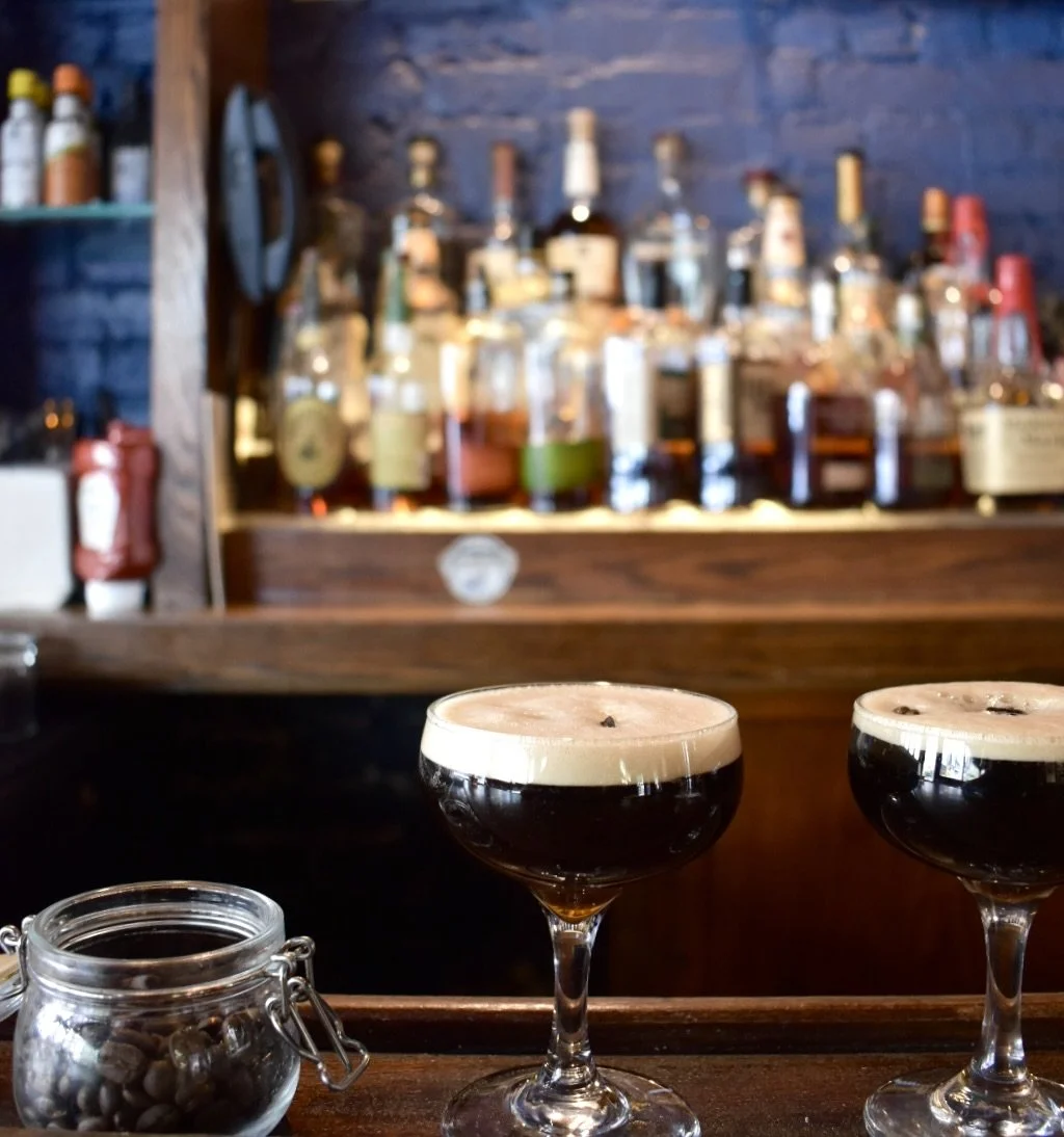 Two glasses of dark beer on a counter with a jar of coffee beans in front, and a backdrop of liquor bottles on shelves in a bar setting.