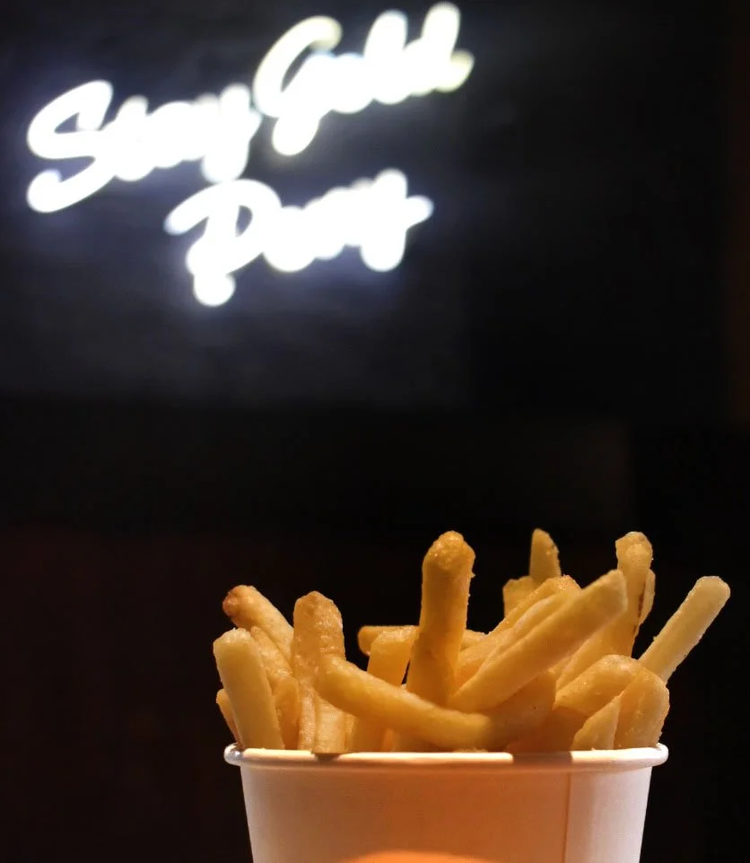 A cup of French fries in front of a dark background with a neon sign that says 'Sweet Gats Puck'.