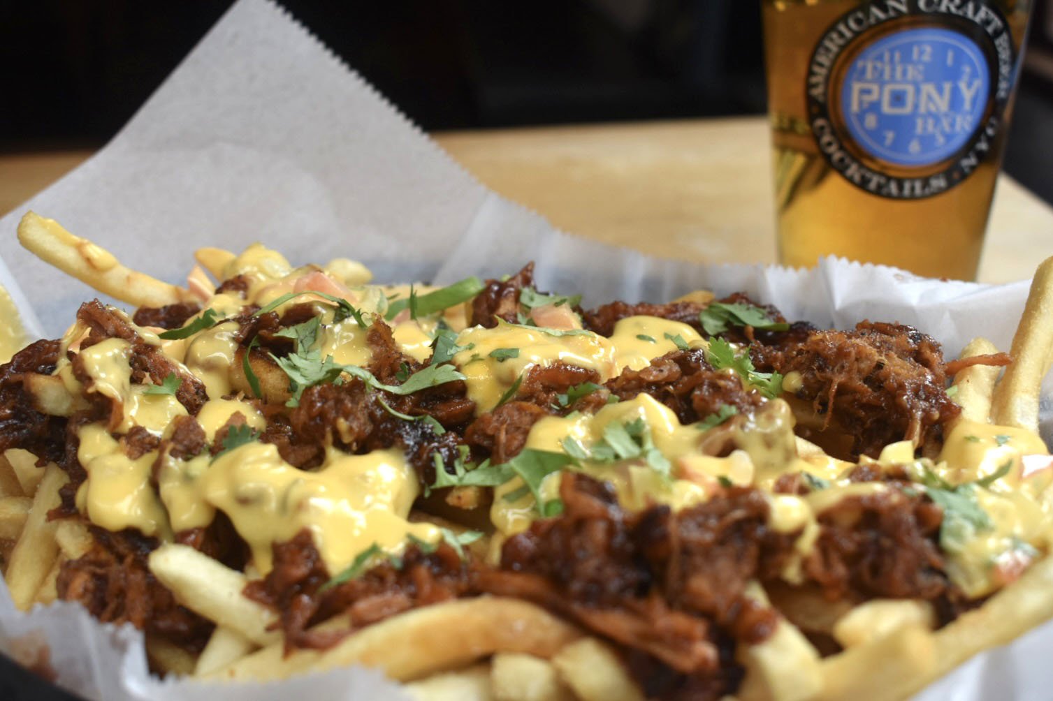 A tray of loaded French fries topped with pulled pork, barbecue sauce, cheese, and fresh cilantro, with a side of lemon and a beverage in the background.