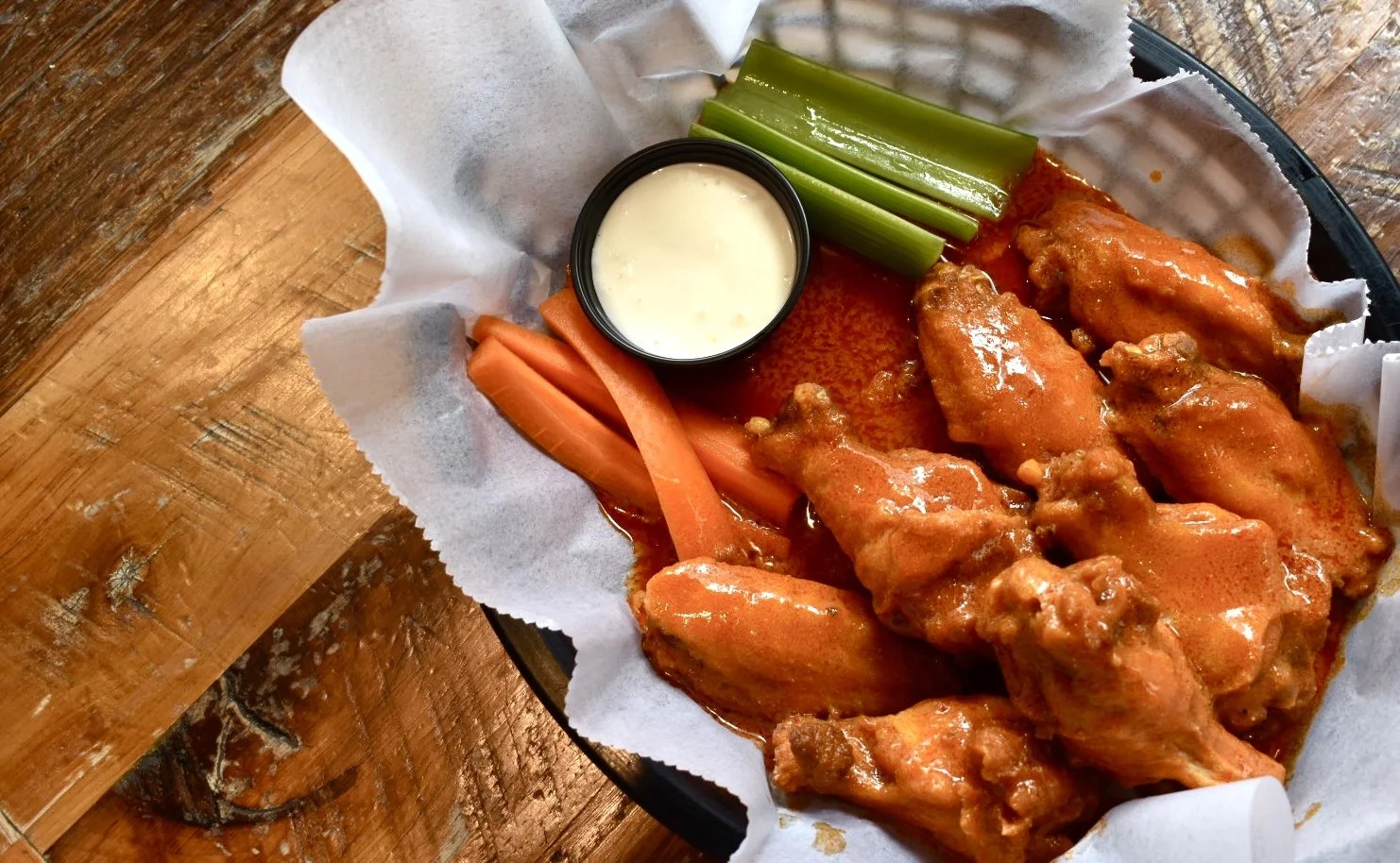 Basket of buffalo chicken wings with celery, carrots, and ranch dipping sauce on a wooden table.