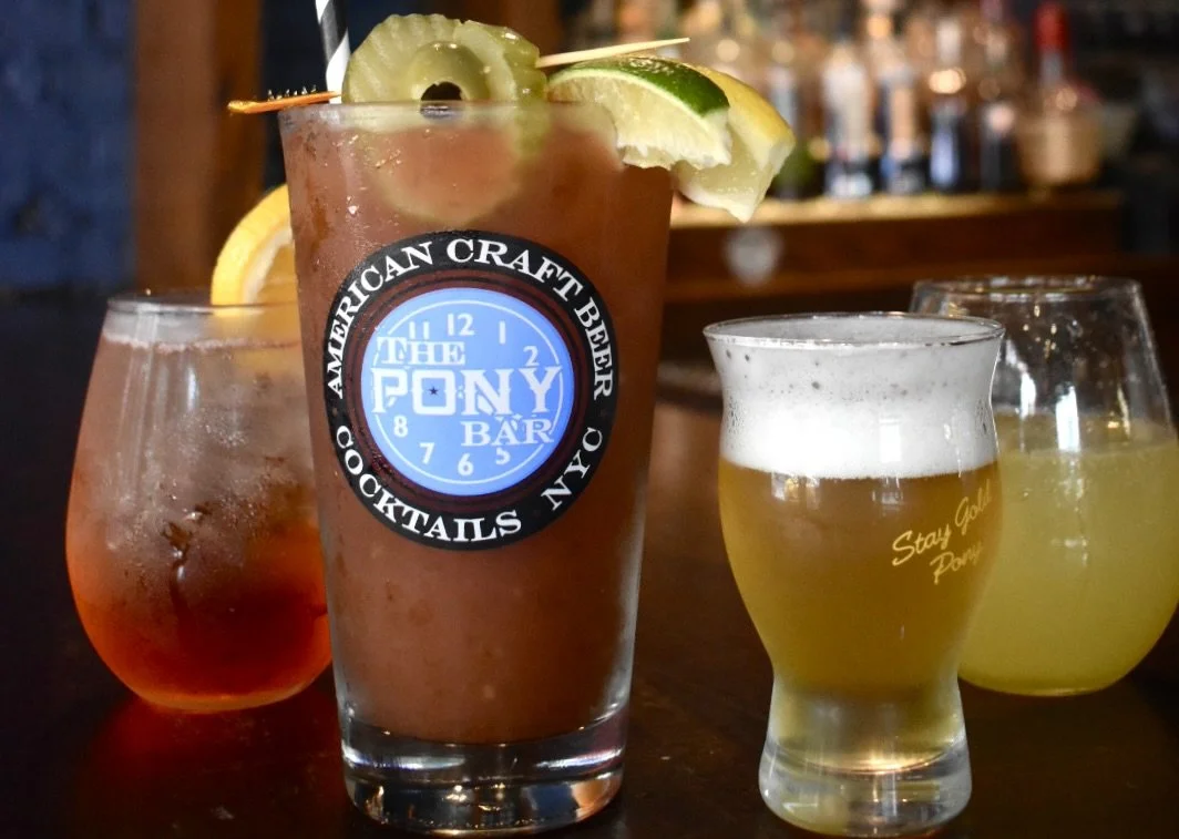 Four cocktails, including a large Bloody Mary with lime and pickle garnish, on a bar counter.