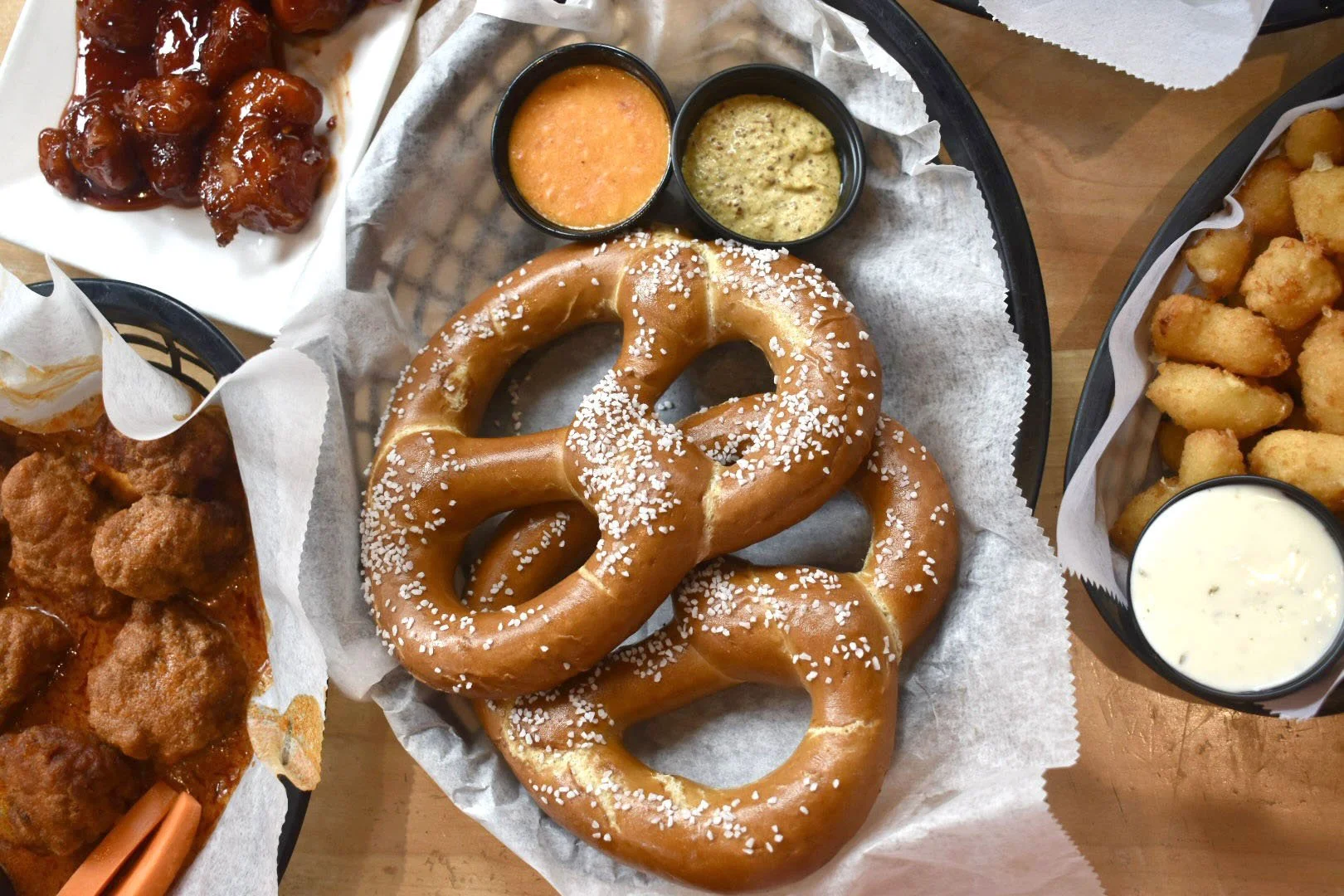 A tray of soft pretzels topped with coarse salt, along with small cups of mustard and cheese sauces, surrounded by baskets of chicken wings in sauce, fried potato nuggets, and sweet and sour chicken.