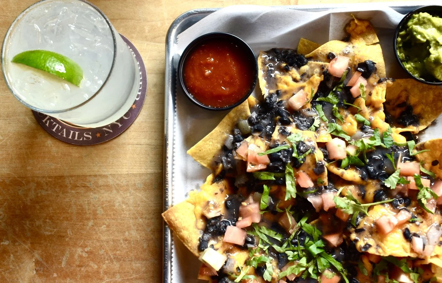A tray with nachos topped with black beans, diced tomatoes, shredded cheese, and cilantro served with a side of guacamole and salsa. Next to the tray, a glass of lime soda with ice and a lime wedge.
