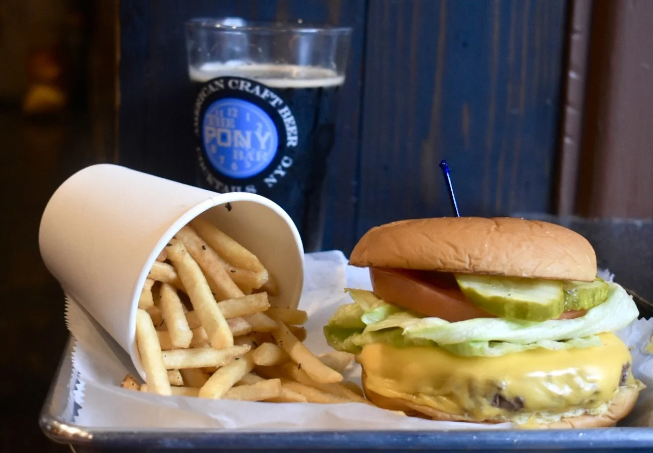 Cheeseburger with lettuce, tomato, pickles, and melted cheese, served with a side of French fries, and a glass of dark soda in the background.