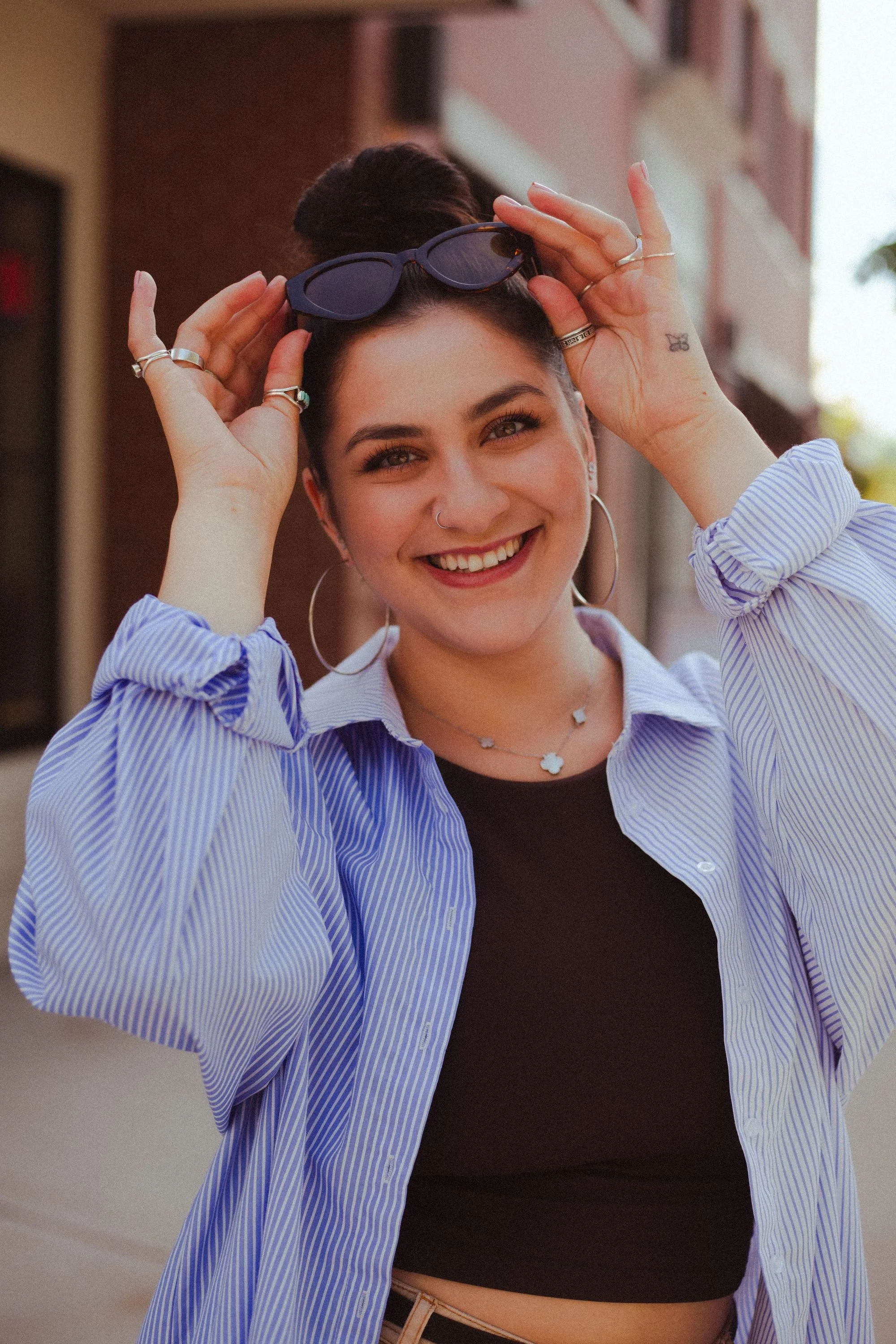 A young woman smiling outdoors, holding sunglasses on her head, wearing a striped blue shirt over a black top, with hoop earrings and jewelry.