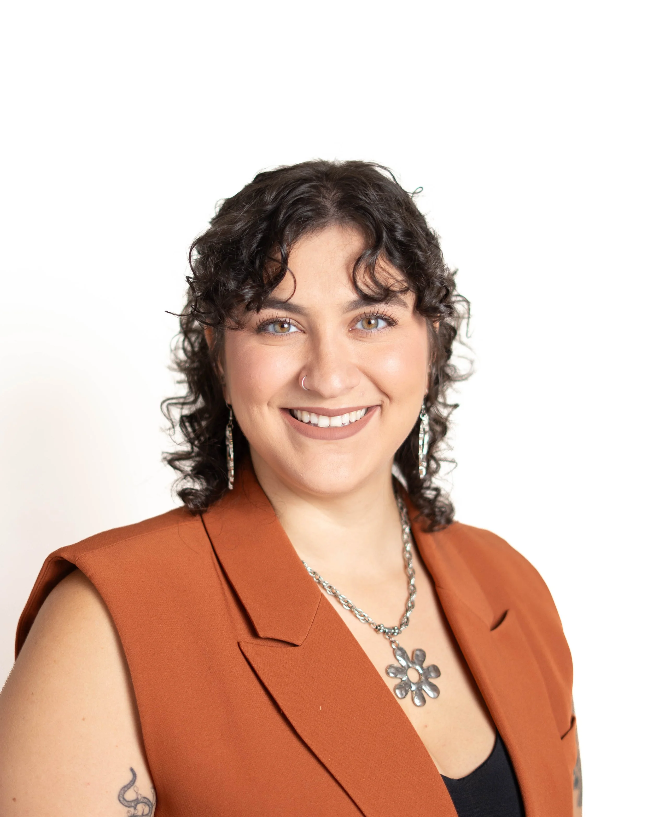 Close-up of a smiling woman with curly brown hair, wearing a rust-colored sleeveless blazer, a black top, a silver floral necklace, and earrings, with a white background.