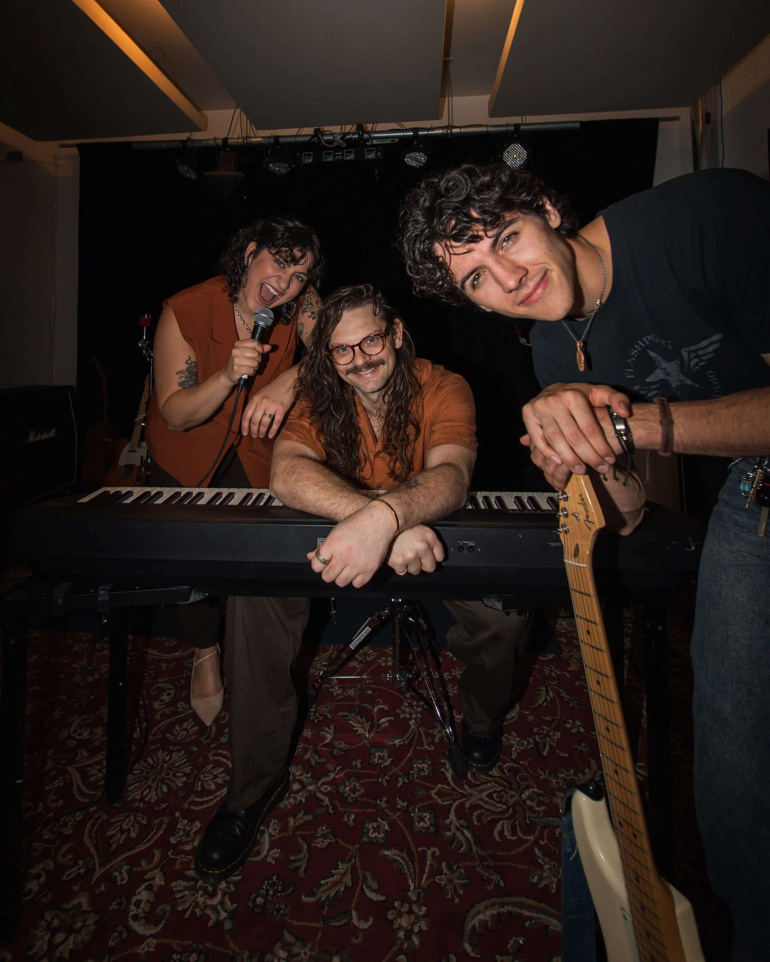 Three musicians in a small performance space, one woman holding a microphone, another man with long hair and glasses, and a third man leaning into the camera, with musical equipment including a keyboard and electric guitar around them.