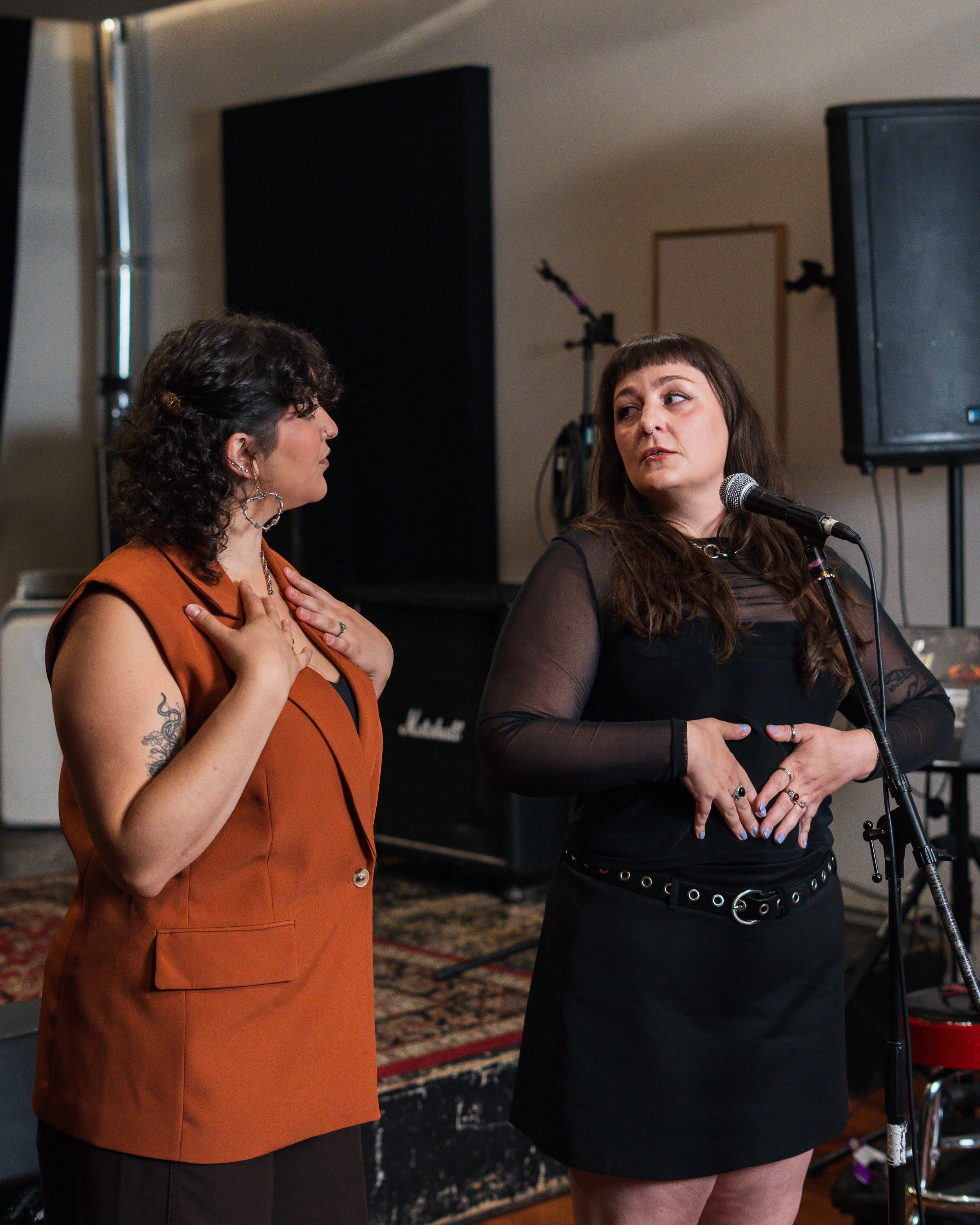 Two women engaged in conversation during an indoor event, one is wearing an orange vest and the other is in a black top and skirt, with microphones and sound equipment visible in the background.