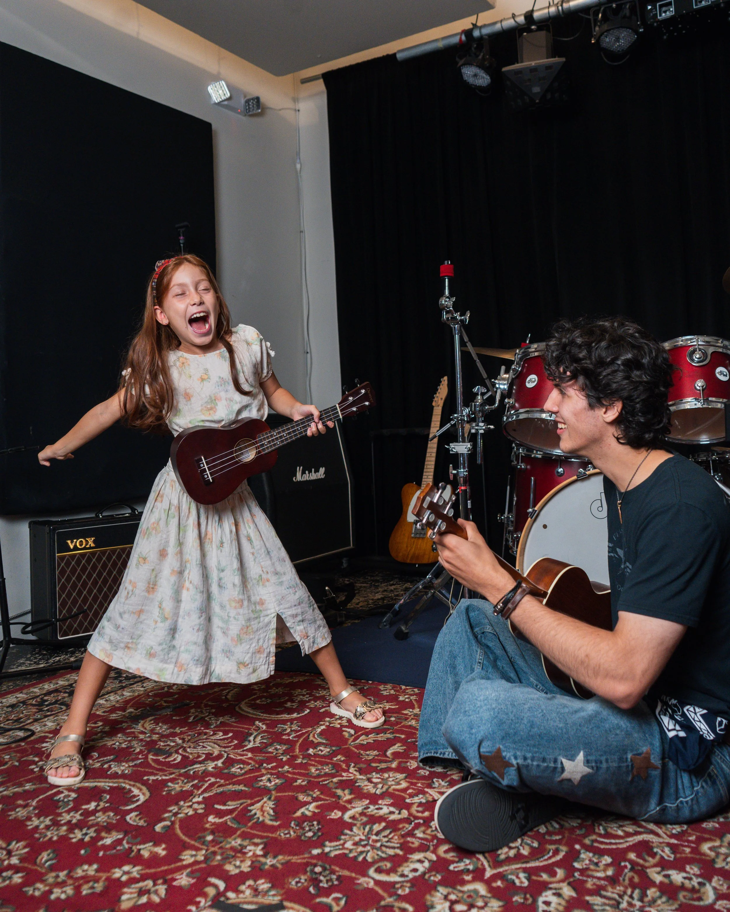 A young girl with long red hair, wearing a floral dress and sandals, passionately singing and playing a ukulele on stage. A man kneeling nearby, smiling and playing an acoustic guitar. The stage has musical instruments, including a drum set and amplifiers, with black curtains and stage lights.