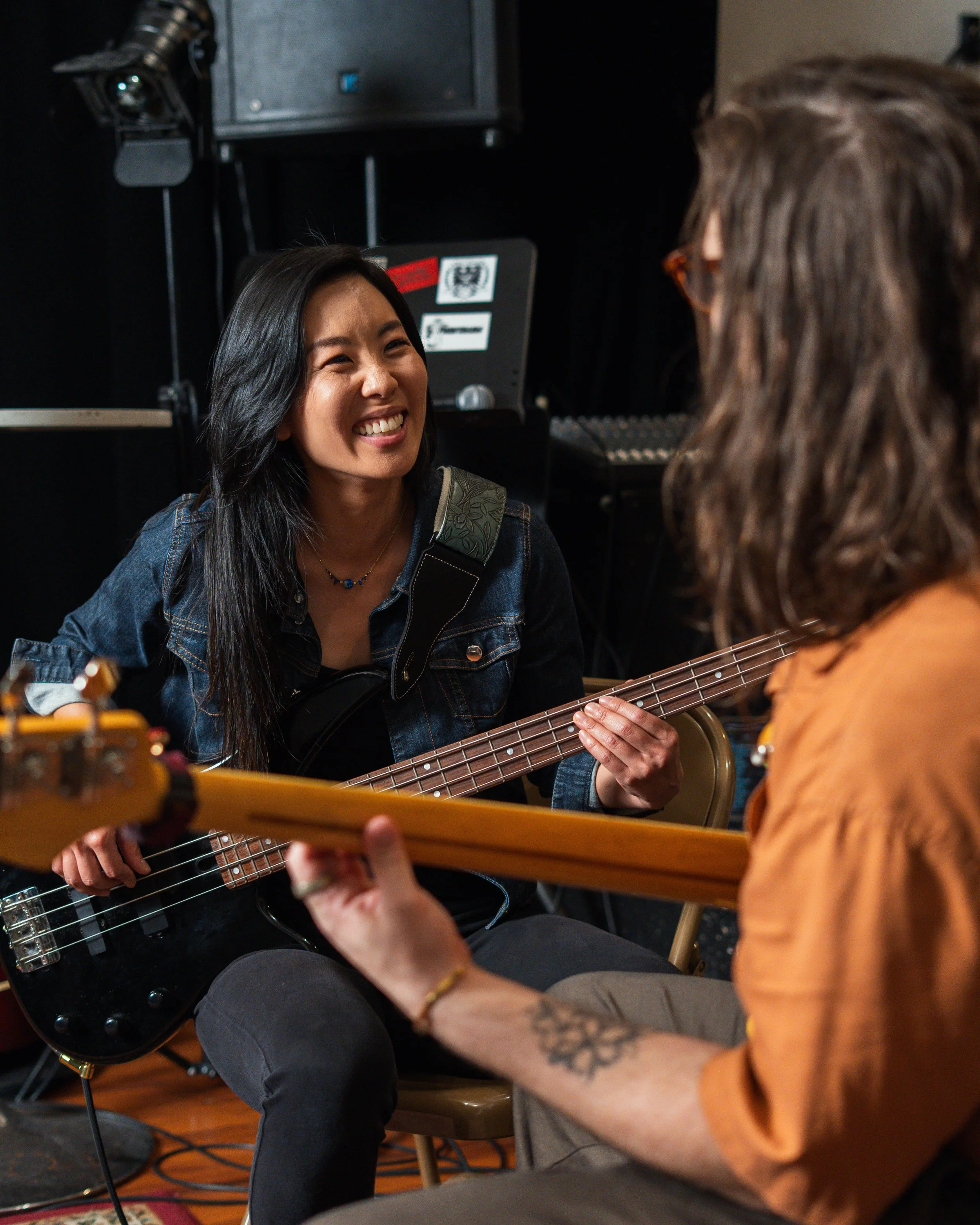 Two people sitting and playing guitars, smiling and engaging in a music session in a room with music equipment in the background.