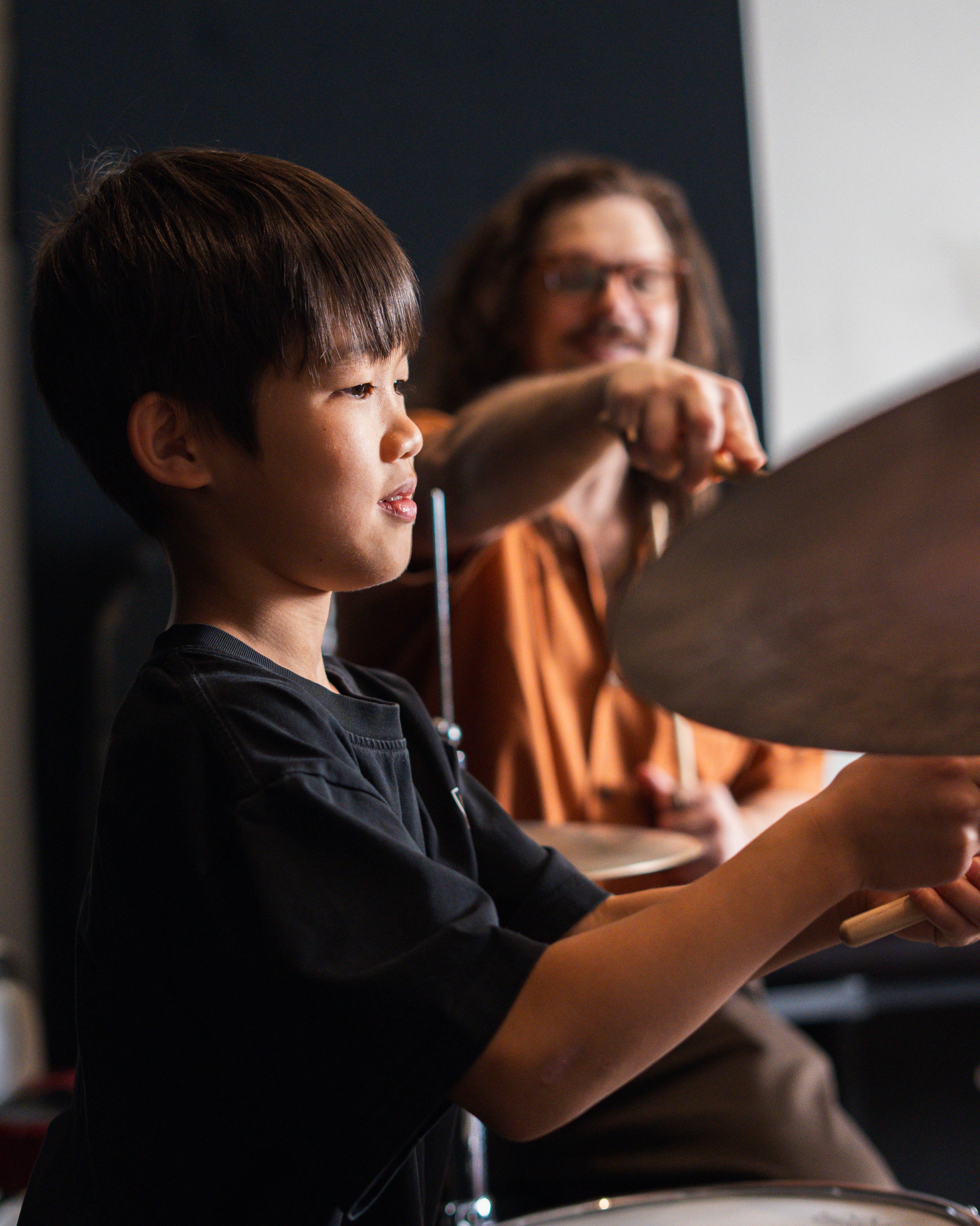 A young boy with dark hair playing drums next to a woman with glasses, in a music studio or practice room.