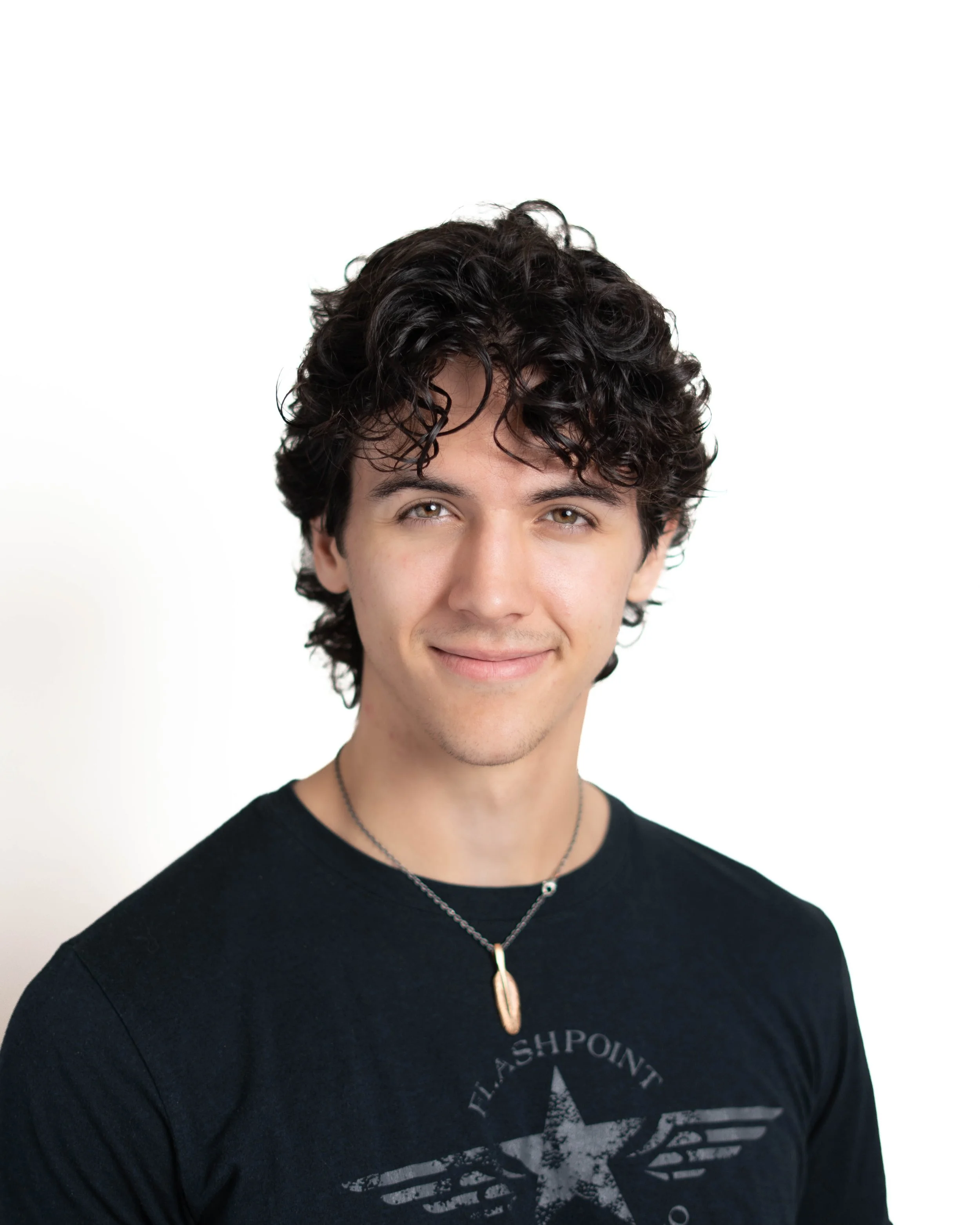 Close-up of a young man with curly dark hair, wearing a black t-shirt with a star and text, and a silver necklace with a pendant, smiling softly against a plain white background.