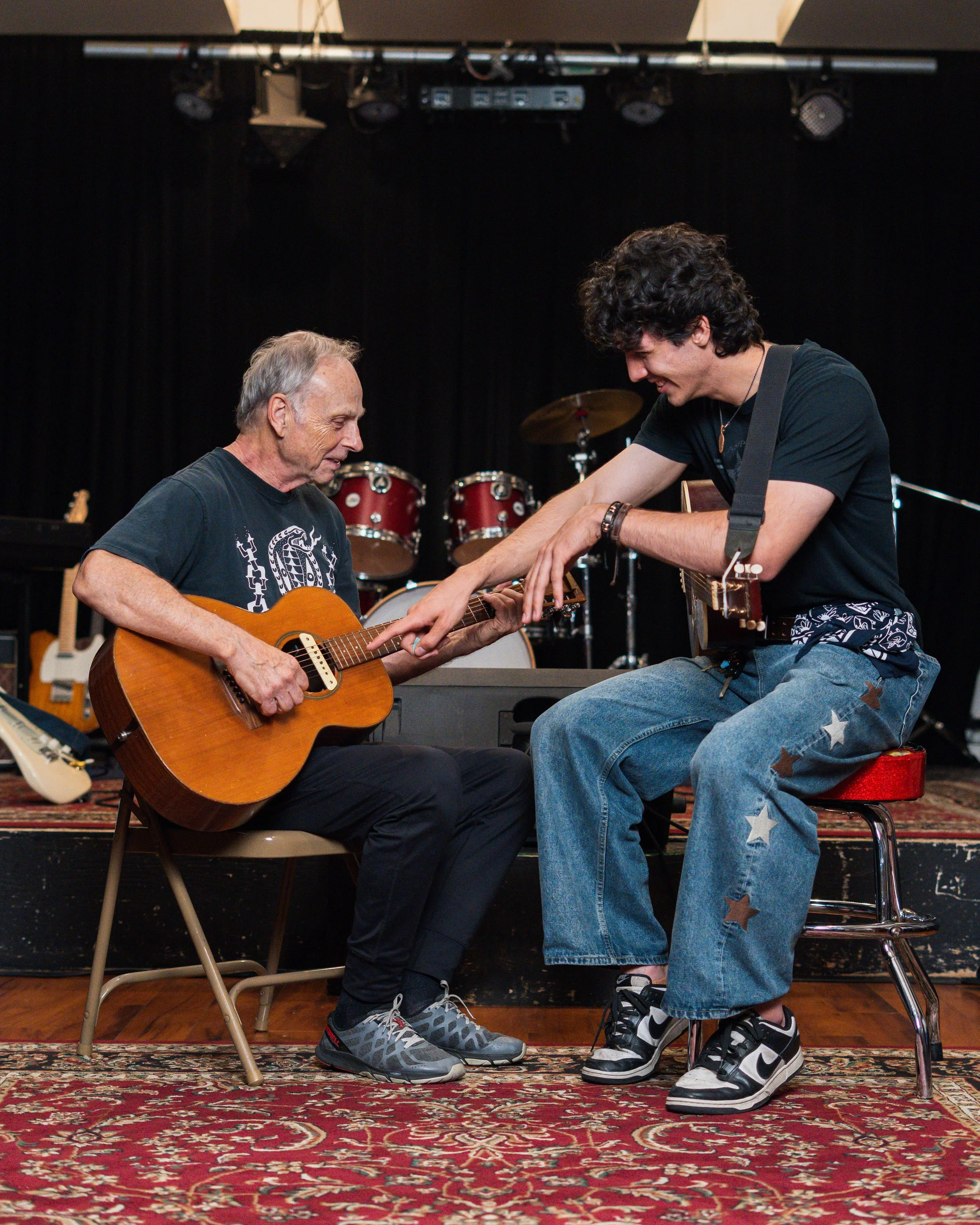 Older man and younger man playing guitar together on stage, smiling, with musical instruments including drums in the background, indoors with a patterned rug.