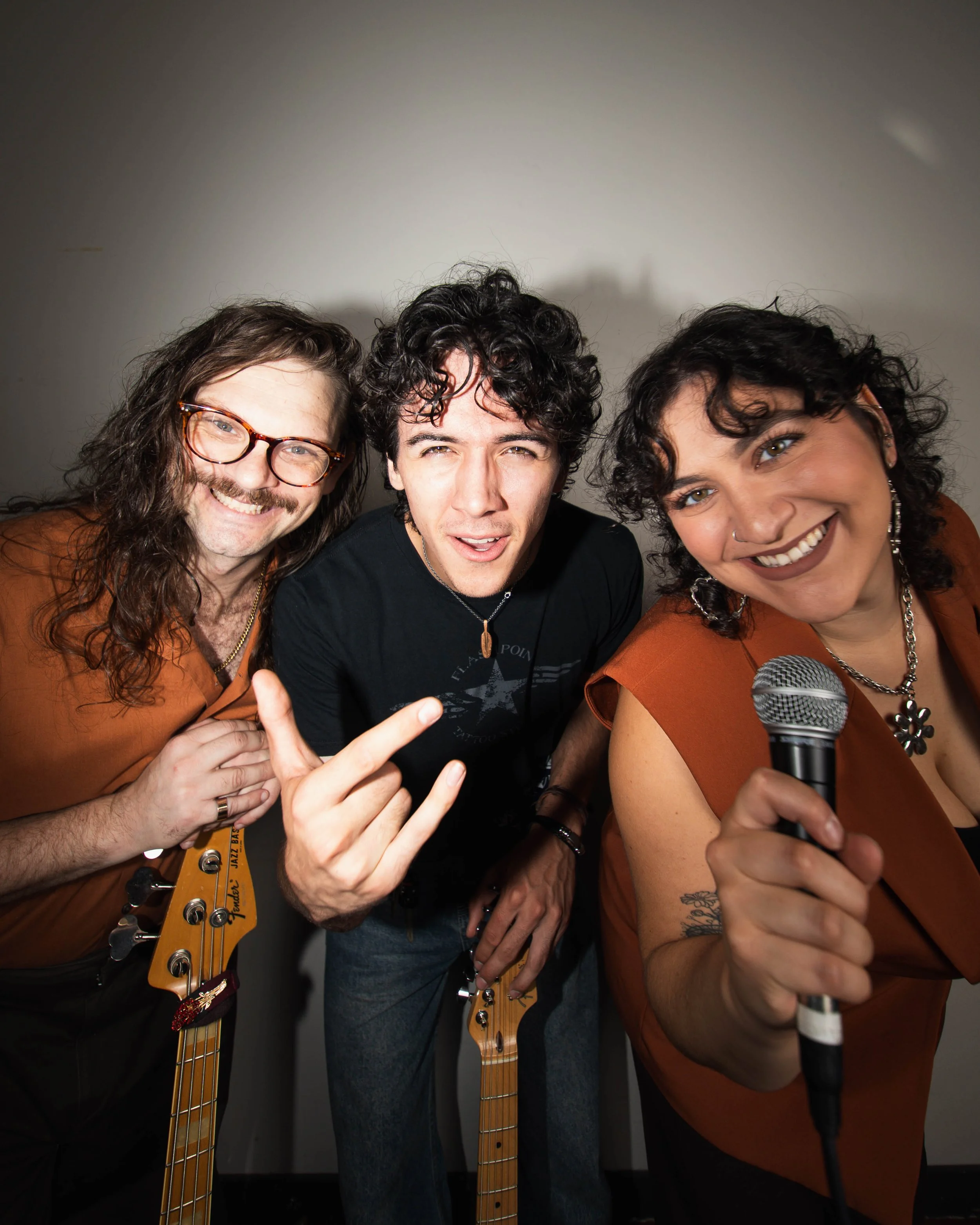 Three musicians close together, two holding guitars and one holding a microphone, smiling and making gestures.