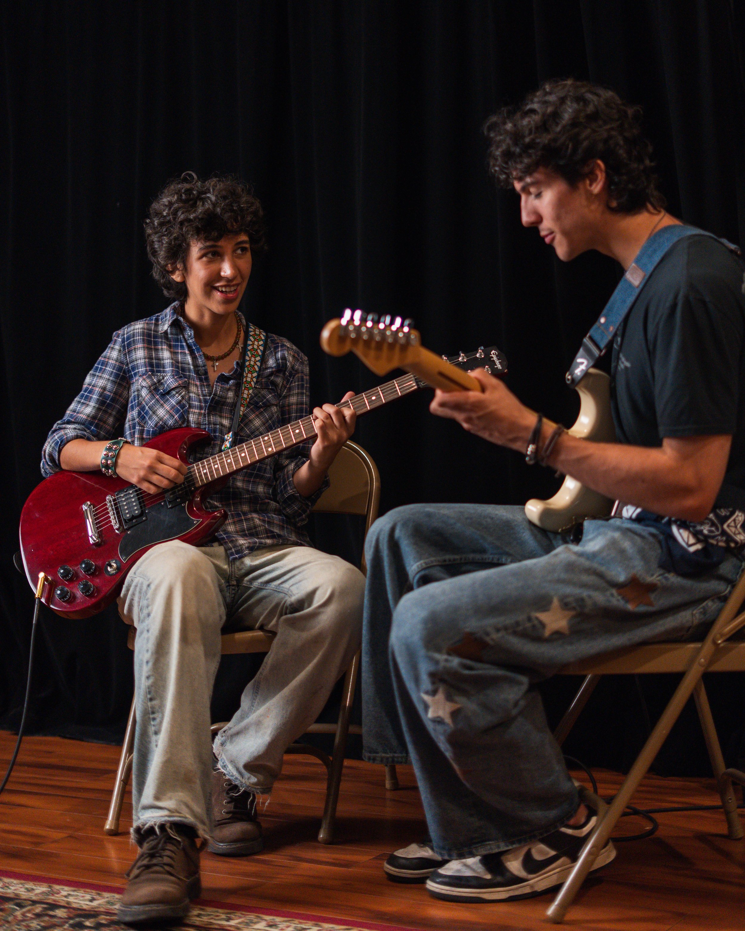 Two young musicians, a woman and a man, sitting on chairs on a wooden stage, playing guitars and engaging with each other. The woman, with curly hair, wears a plaid shirt, beige pants, and a beaded bracelet. The man, with curly hair, wears a dark t-shirt and jeans, and is looking down at his guitar. The background is black, and part of a patterned rug is visible on the floor.