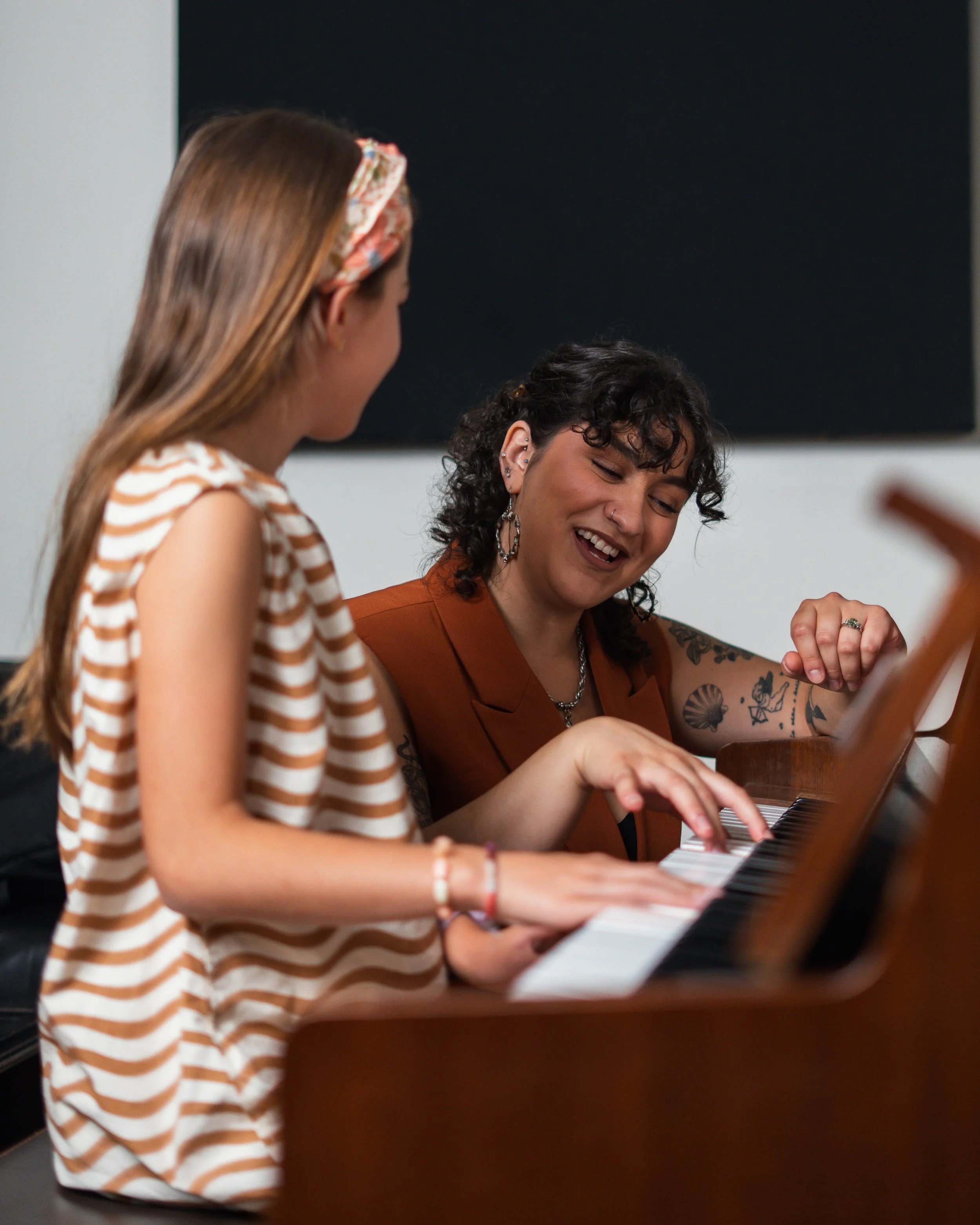 Two women sitting at a piano, smiling and engaging with each other, in an indoor setting.