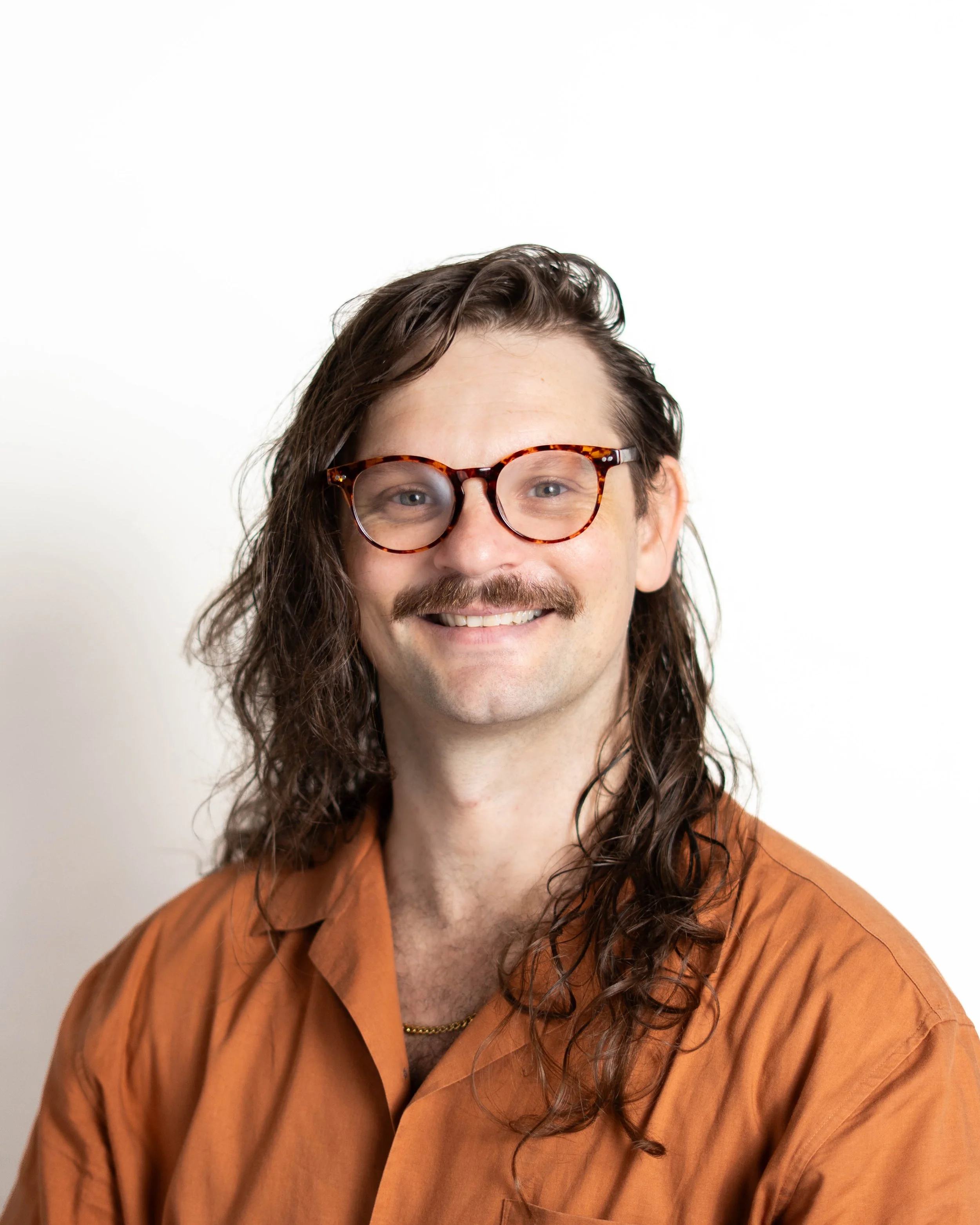 A person with long curly hair, wearing tortoiseshell glasses, smiling, dressed in an orange-brown shirt, against a plain white background.